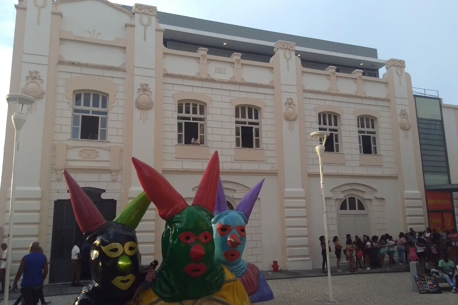 The Casa do Carnaval from the square outside: three giant painted Carnival-mask sculptures in green, black and blue set out in front of the museum’s pale 1911 facade, with a line of visitors along the pavement by the entrance
