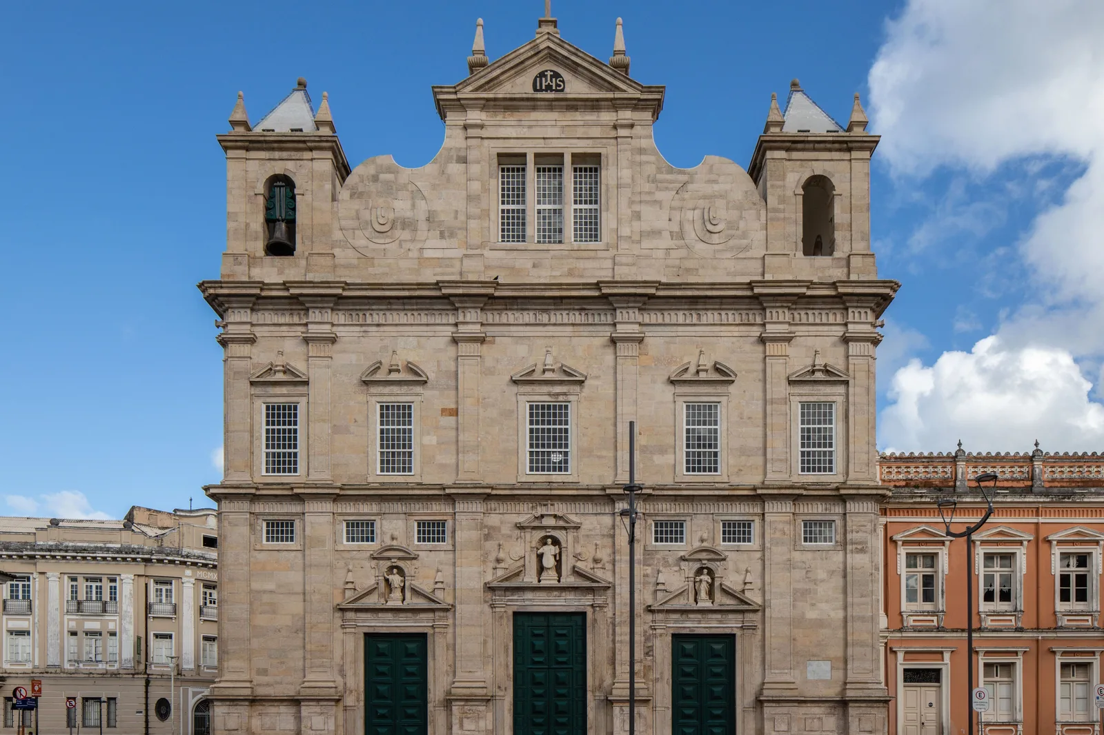 The pale limestone facade of the Catedral Basílica filling the frame from across the Terreiro de Jesus, three green doors below tiers of pilasters and grilled windows, with the colonial houses of the square either side of it