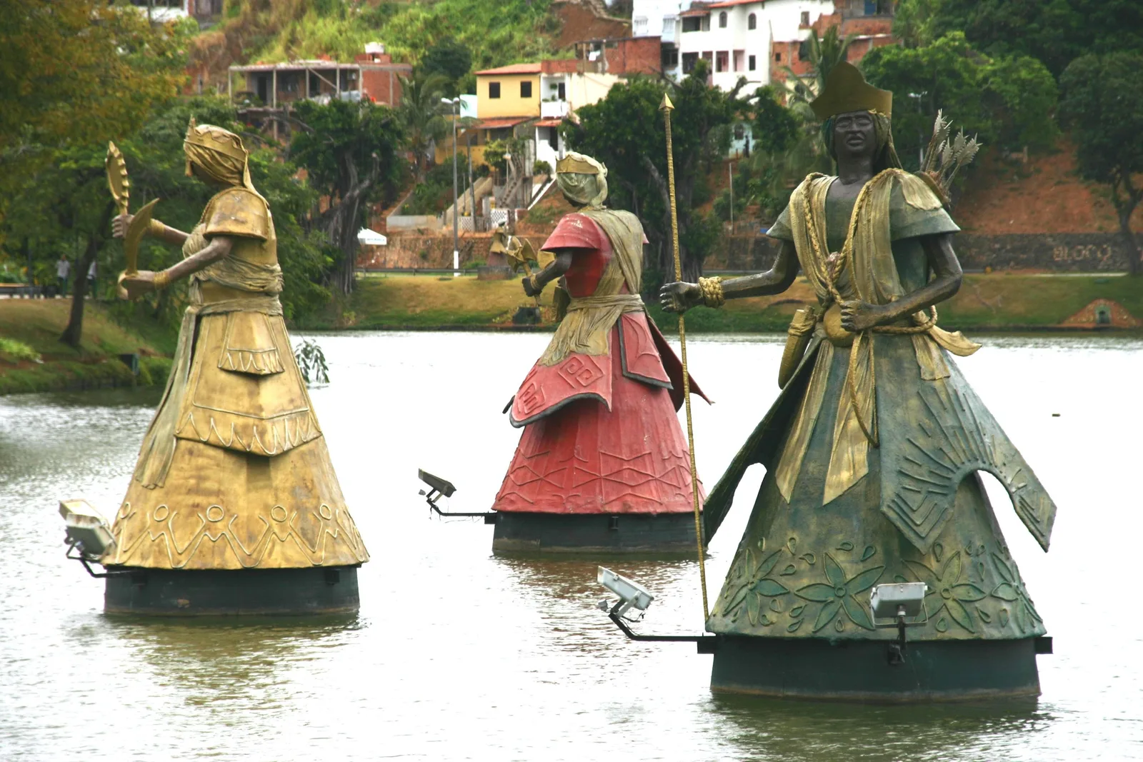 Three of the orixá sculptures standing out on the water of the Dique do Tororó, robed figures in gold, red and green holding their emblems, with the houses of the hillside neighborhood rising behind the lake