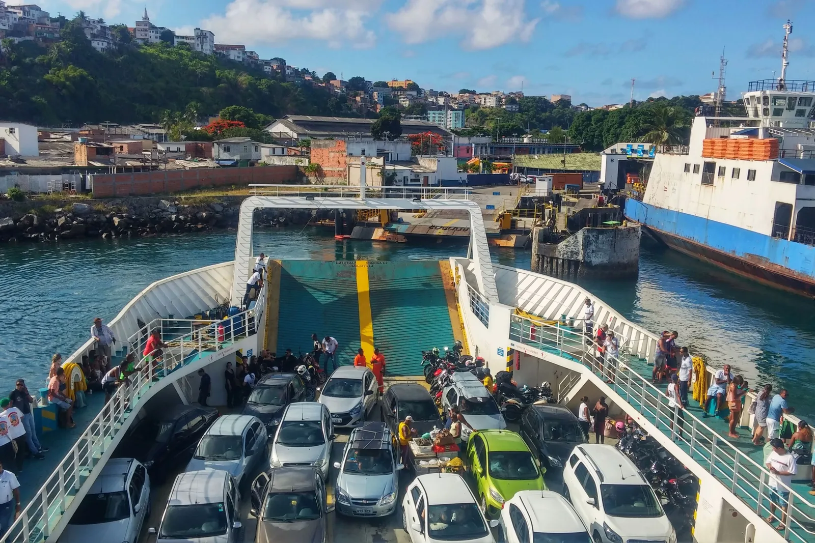 The car deck of a ferry at the São Joaquim terminal in Salvador, cars and motorbikes packed in bumper to bumper, passengers standing along the rails and another ferry tied up alongside with the hillside of the city behind