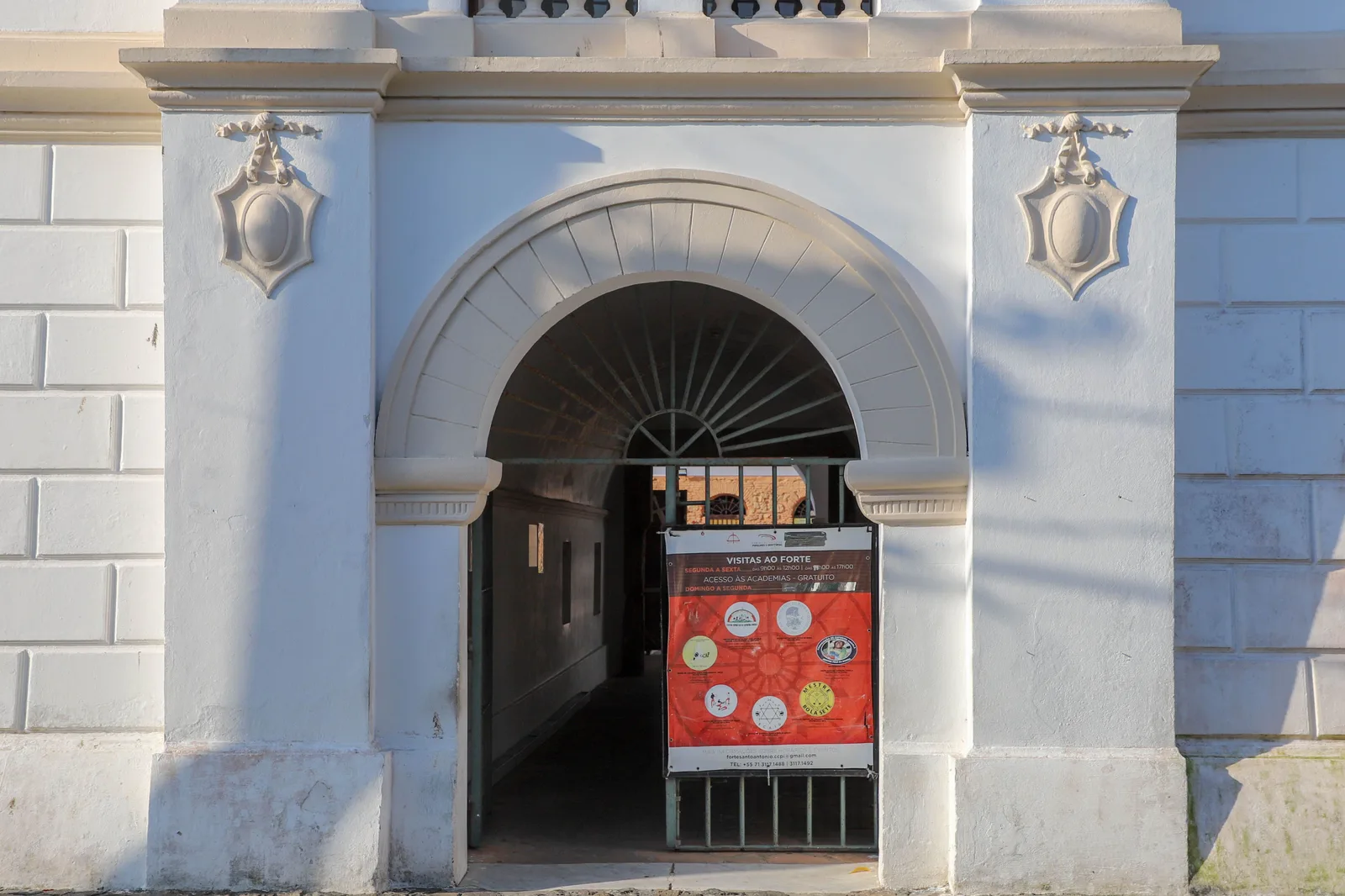 The whitewashed arched gateway of the Forte de Santo Antônio Além do Carmo in low sun, with a red board standing inside the arch that lists the capoeira academies working in the fort and free access to them