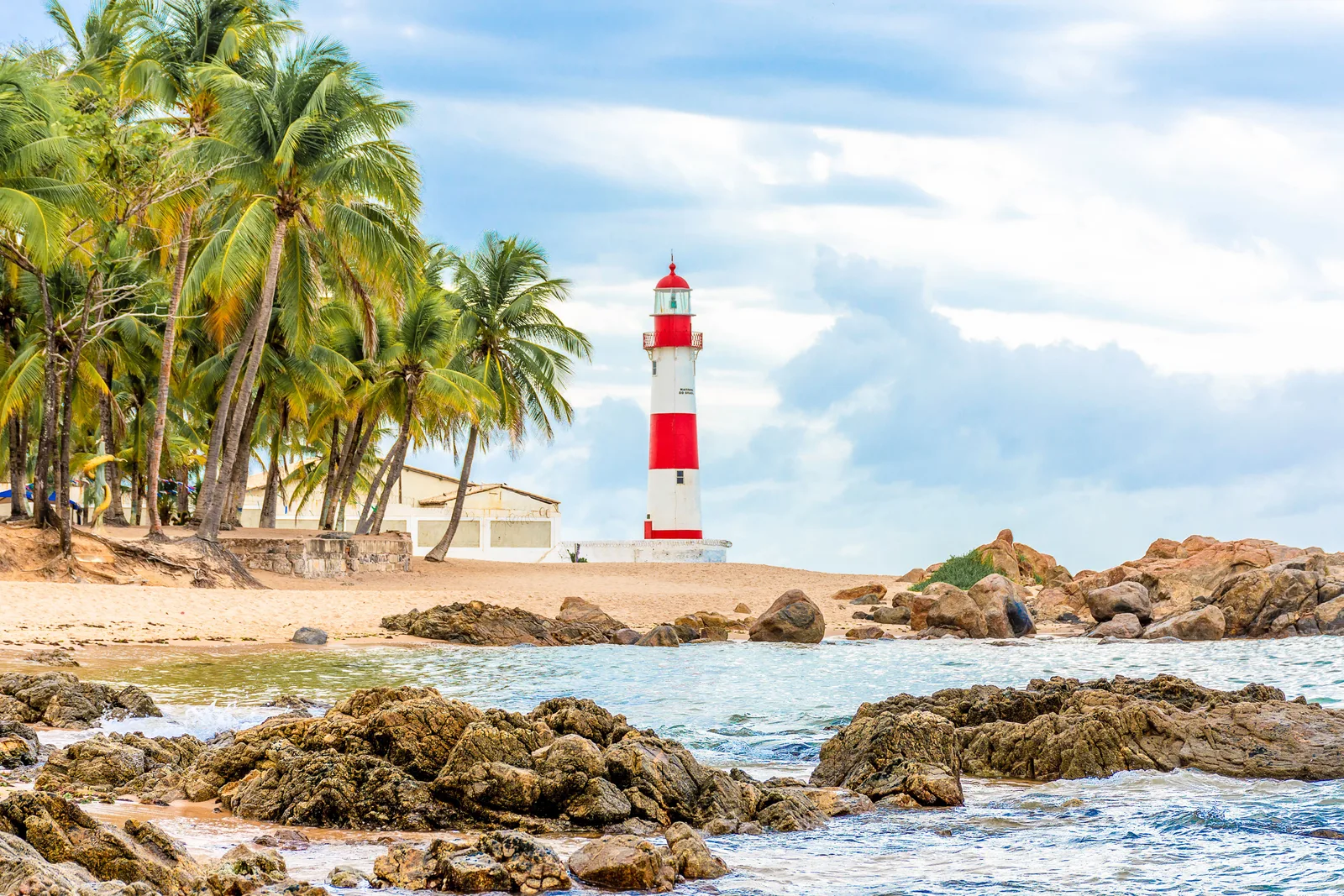 The red-and-white Itapuã lighthouse standing behind a line of coconut palms on the sand, with dark reef rock breaking the surface of the shallow water in the foreground