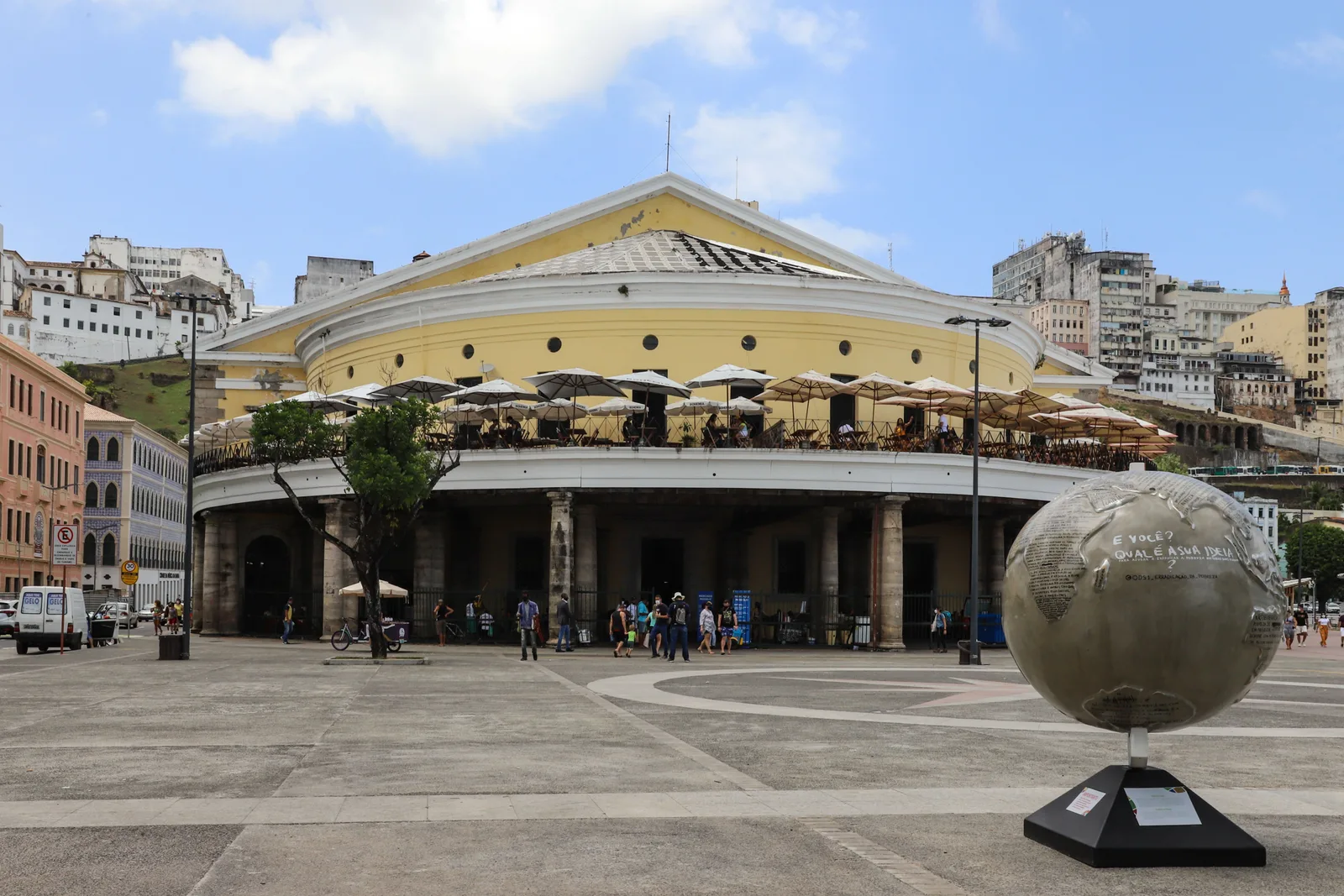 The Mercado Modelo from the plaza in front of it: the old yellow customs house with white umbrellas shading the restaurant terrace on its upper floor, shoppers going in and out under the stone arcade below, and the cliff of the upper city rising behind