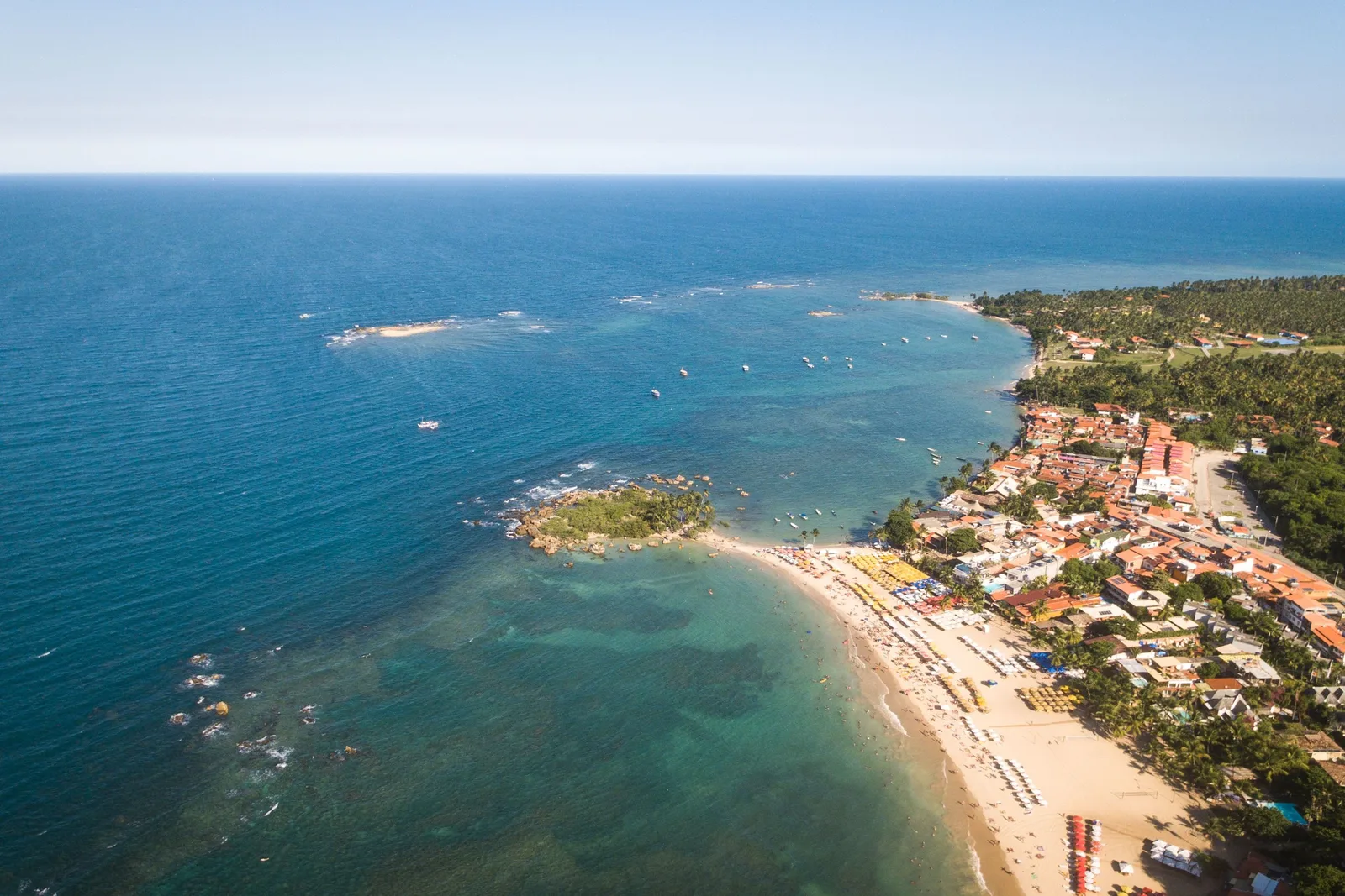 Morro de São Paulo from the air: a long arc of pale sand lined with beach umbrellas, low red-roofed pousadas and palms packed in right behind it, and reef showing dark through turquoise water offshore