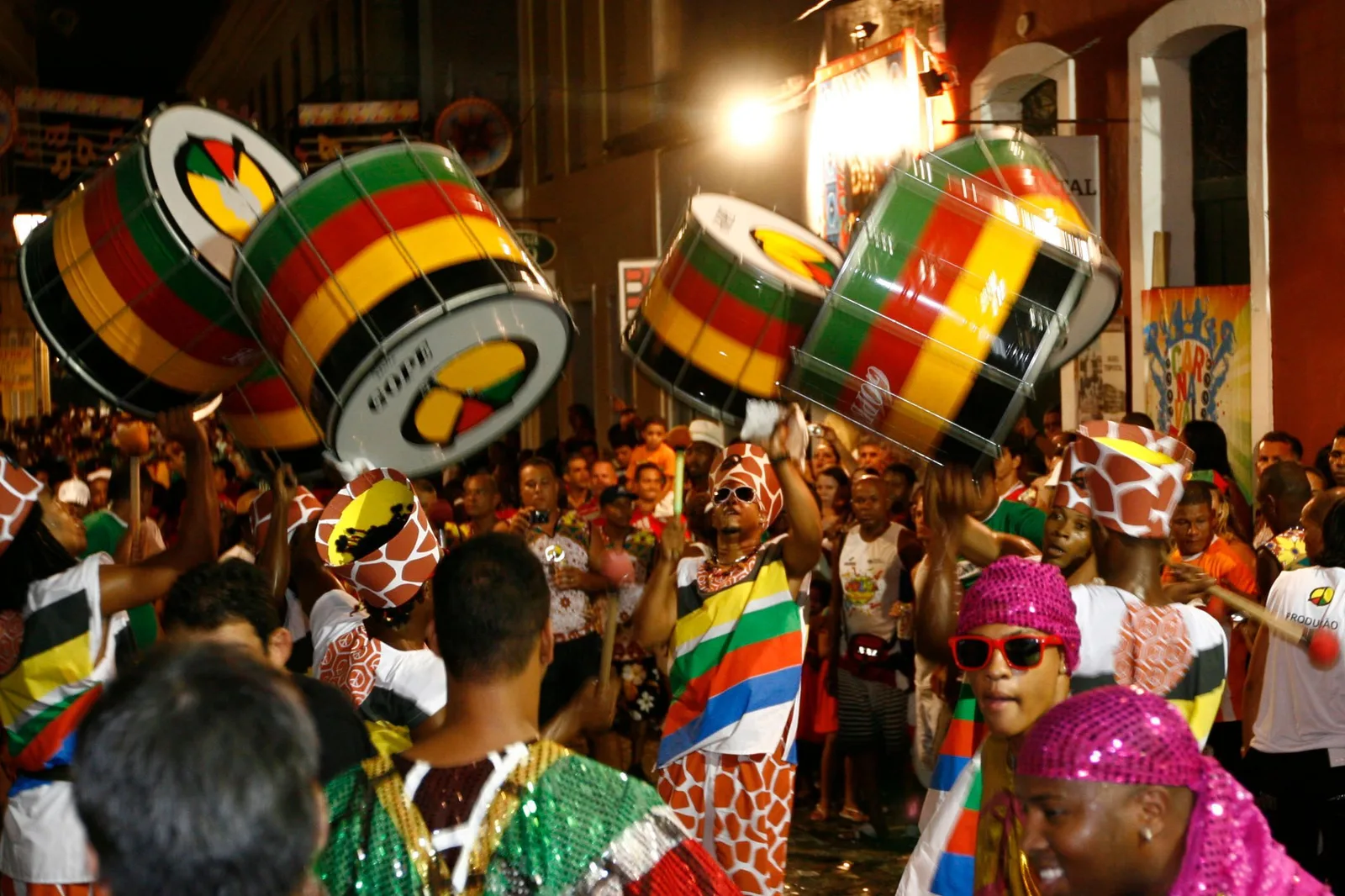 Olodum drummers moving off through a Salvador street at night, big surdos painted in green, yellow and red held up over their shoulders, players in printed headwraps and a crowd filling the road behind them
