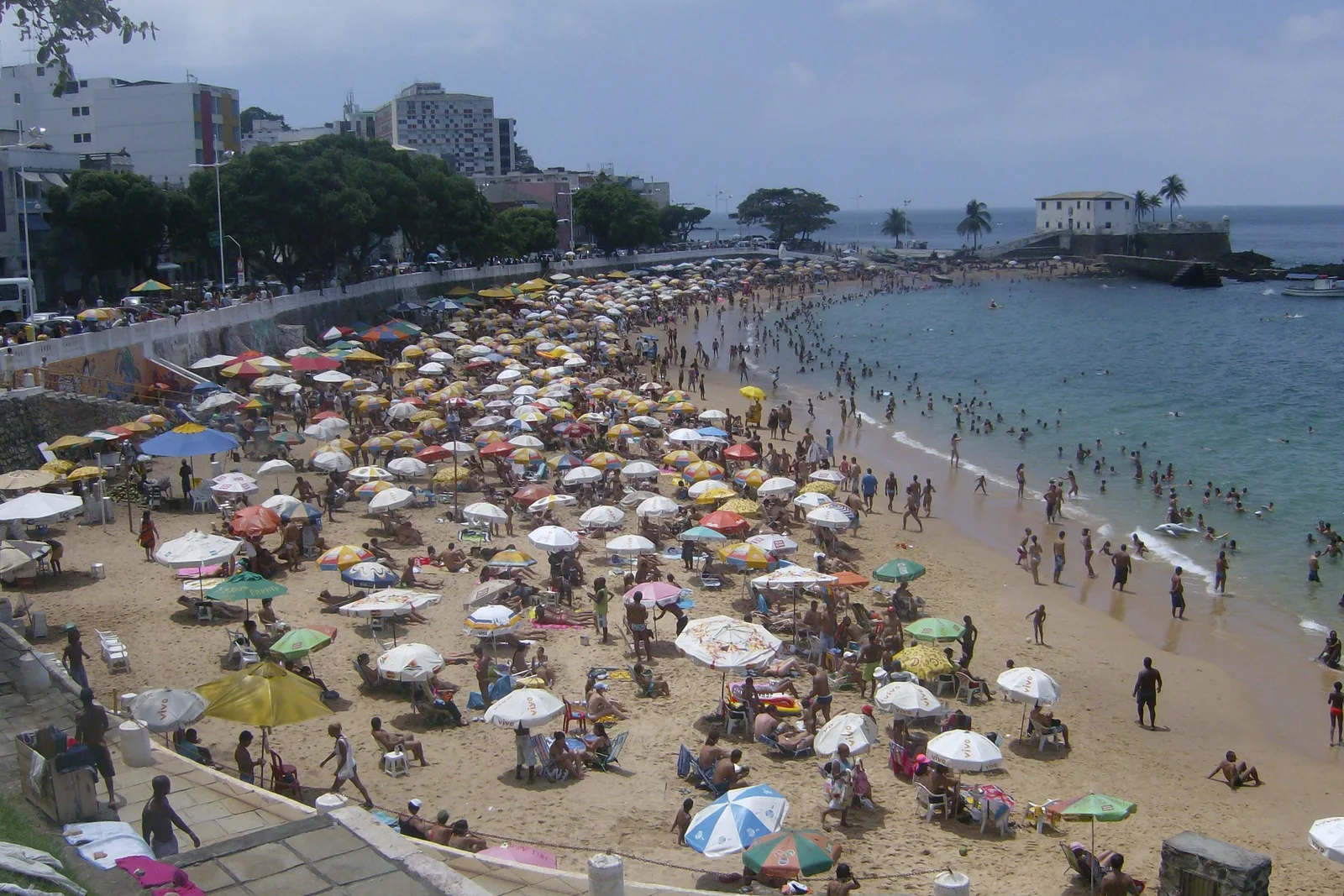 Porto da Barra on a busy afternoon: a curve of sand packed with colored umbrellas, swimmers standing in flat bay water, and the low stone fort closing the far end of the beach