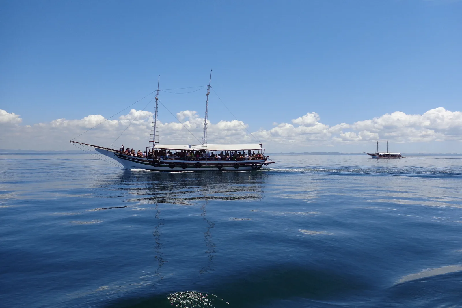 A wooden two-masted schooner under way on the flat blue water of the Bay of All Saints, passengers packed along its awning deck, with a second schooner small on the horizon behind