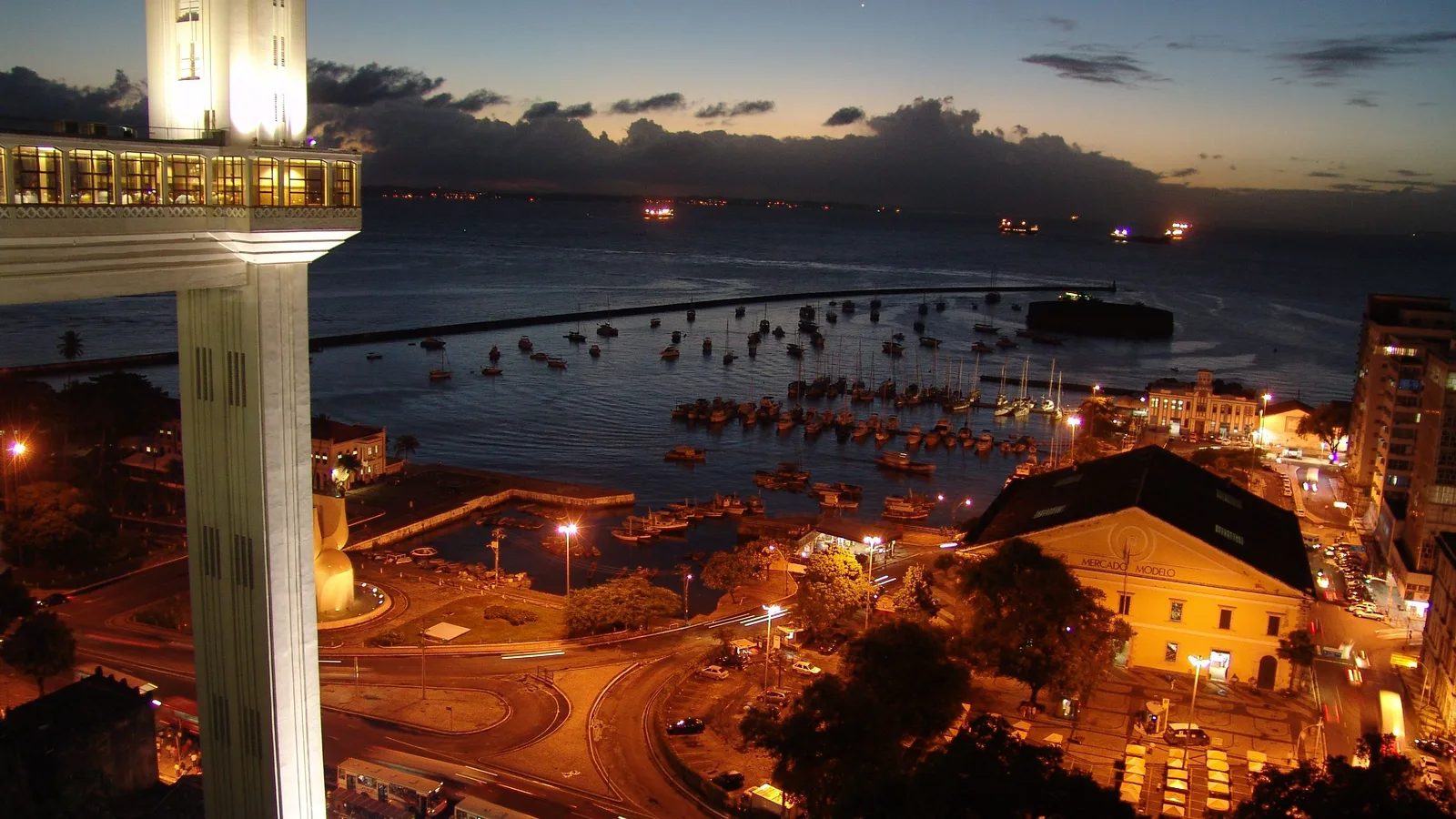 Salvador at dusk from the top of the cliff: the floodlit Elevador Lacerda standing in the foreground, the Mercado Modelo and a marina full of moored boats below it, and the Bay of All Saints running out dark to the horizon