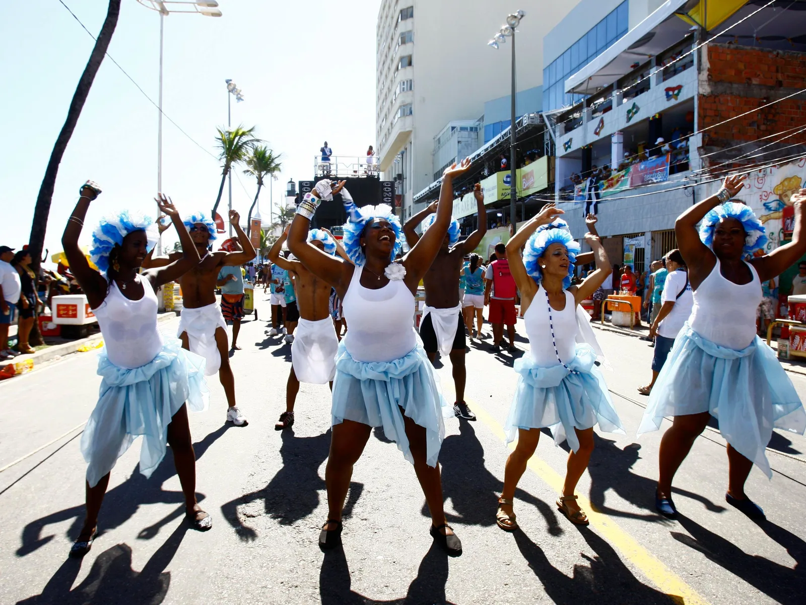 Dancers in white vests and pale blue skirts leading a bloco up the middle of Salvador’s Barra–Ondina Carnival circuit in hard daylight, a sound truck and the tiered spectator boxes lining the avenue behind them