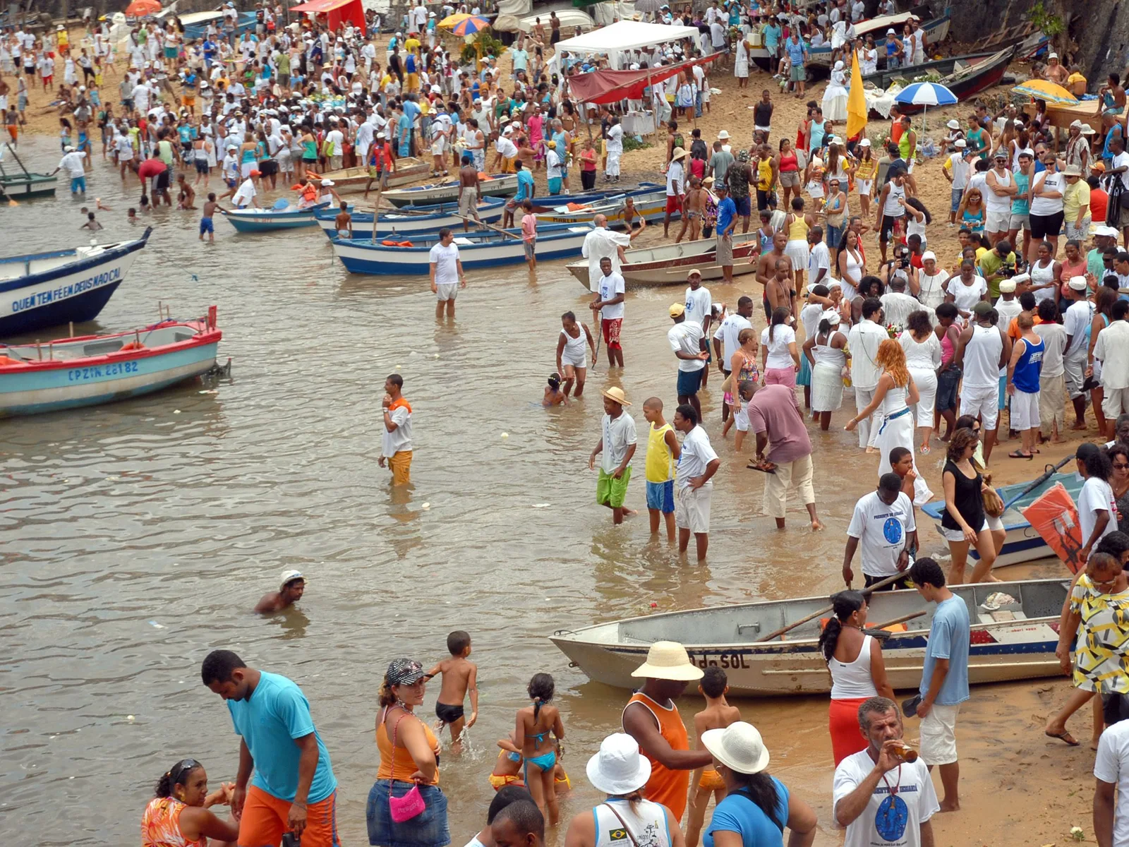 The beach at Rio Vermelho on the day of the Festa de Iemanjá: wooden fishing boats drawn up at the water’s edge, people wading out to them, and the sand behind packed with a crowd mostly dressed in white