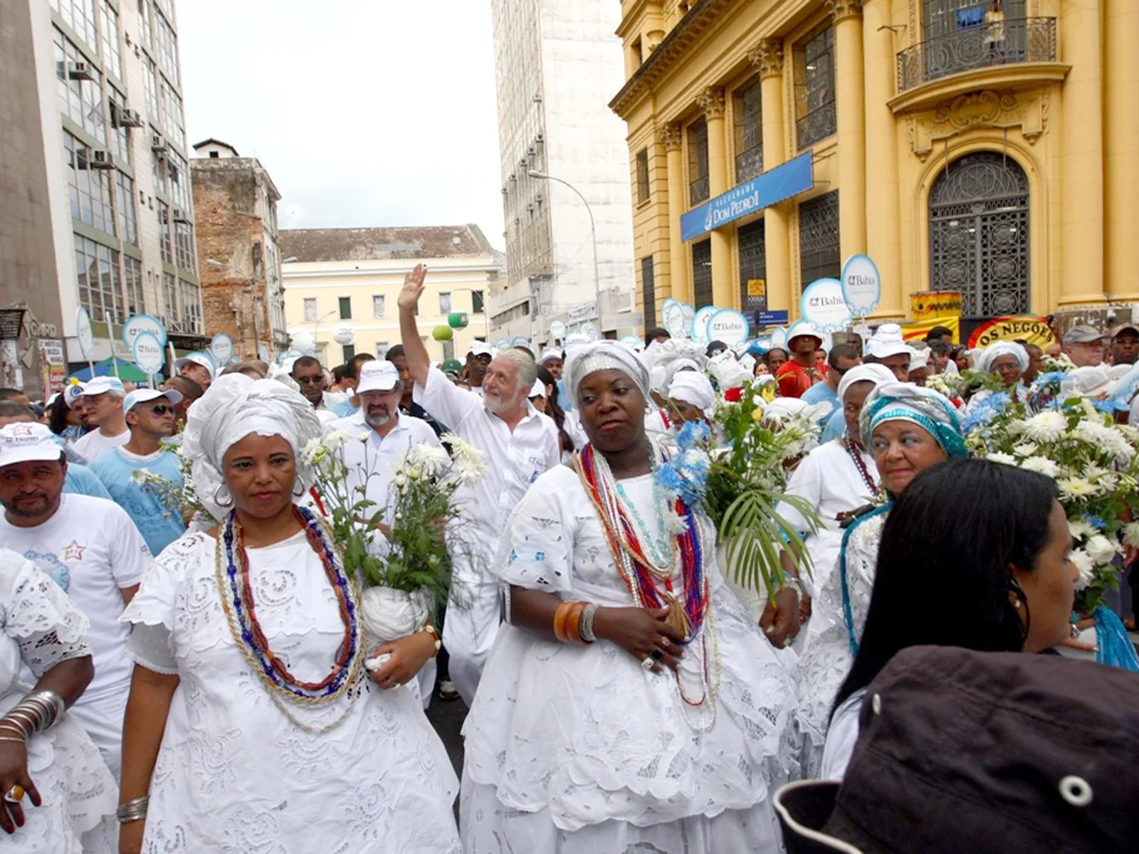 Baianas in white lace dresses, headwraps and layered beads carrying armfuls of white flowers through the Lavagem do Bonfim procession, the crowd dressed in white filling the street behind them
