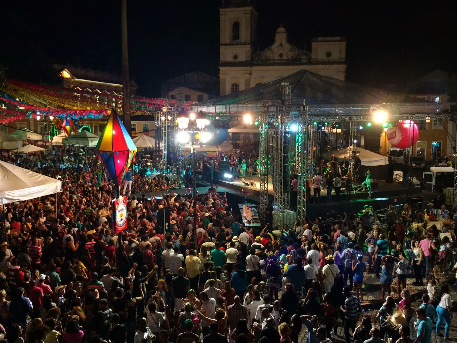 São João night on the Terreiro de Jesus: strings of paper bunting and a big striped balloon lantern over the square, a lit stage at the far side, and a crowd filling the cobbles in front of the cathedral