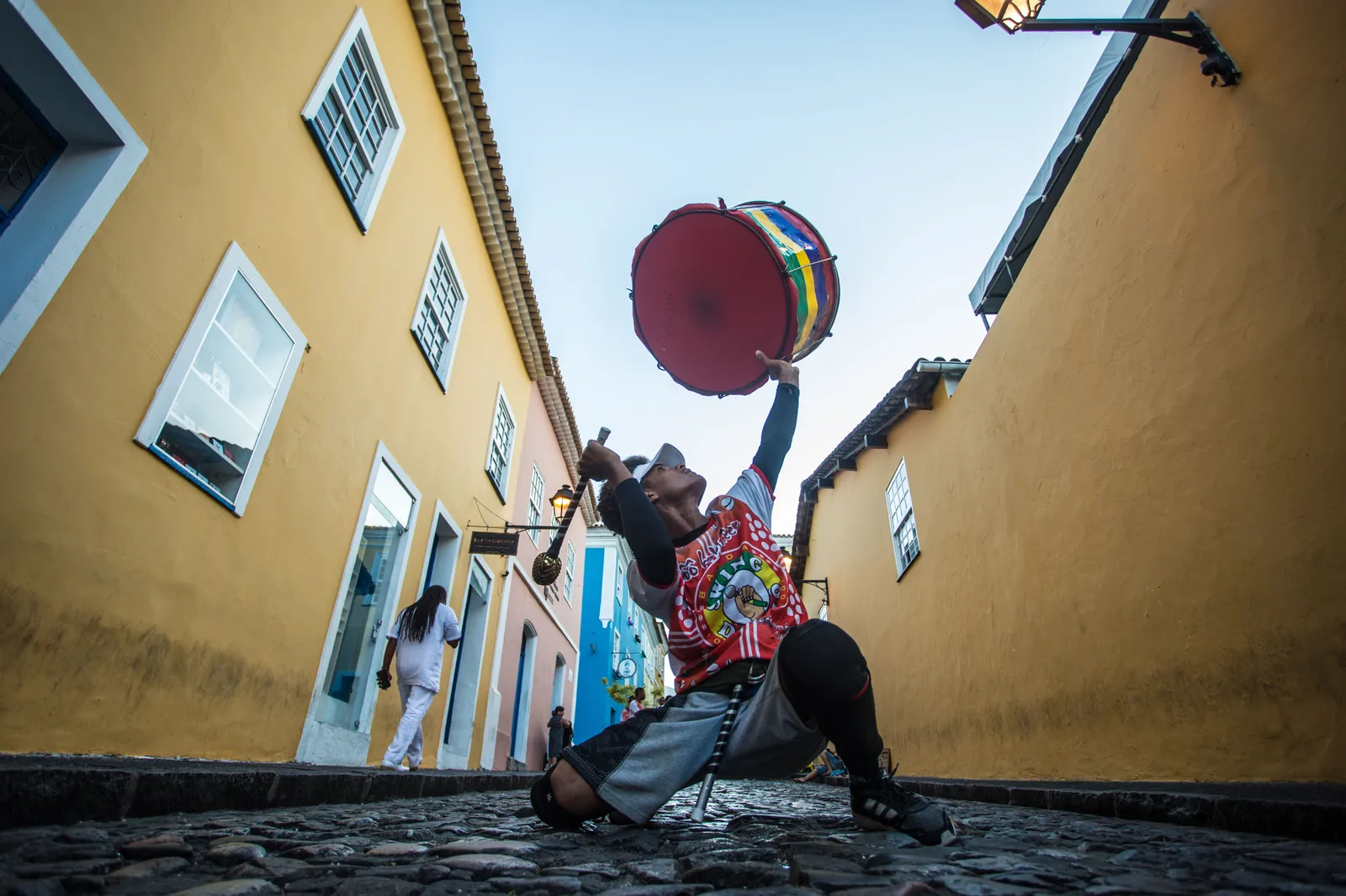 A drummer leaping in mid-air with his drum in a narrow Pelourinho street of yellow colonial houses