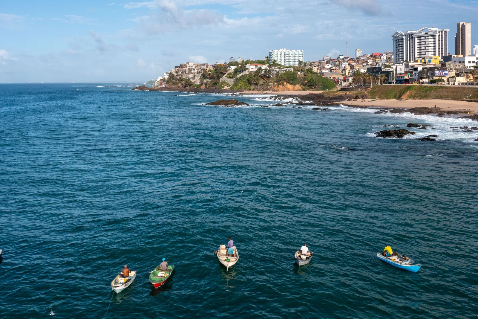 Fishing canoes on deep blue water off Rio Vermelho, seen from above, the neighborhood’s rocky shore and houses behind