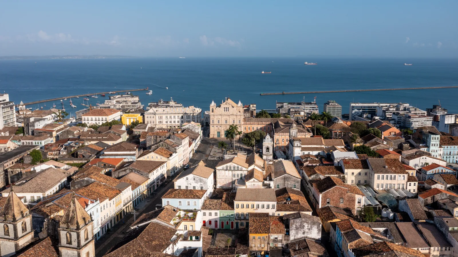 Salvador’s historic center from the air: red-tiled colonial rooftops and church towers packed along the clifftop, the pale facade of the Catedral Basílica on the Terreiro de Jesus in the middle of them, and the Bay of All Saints with the port and anchored ships stretching out behind