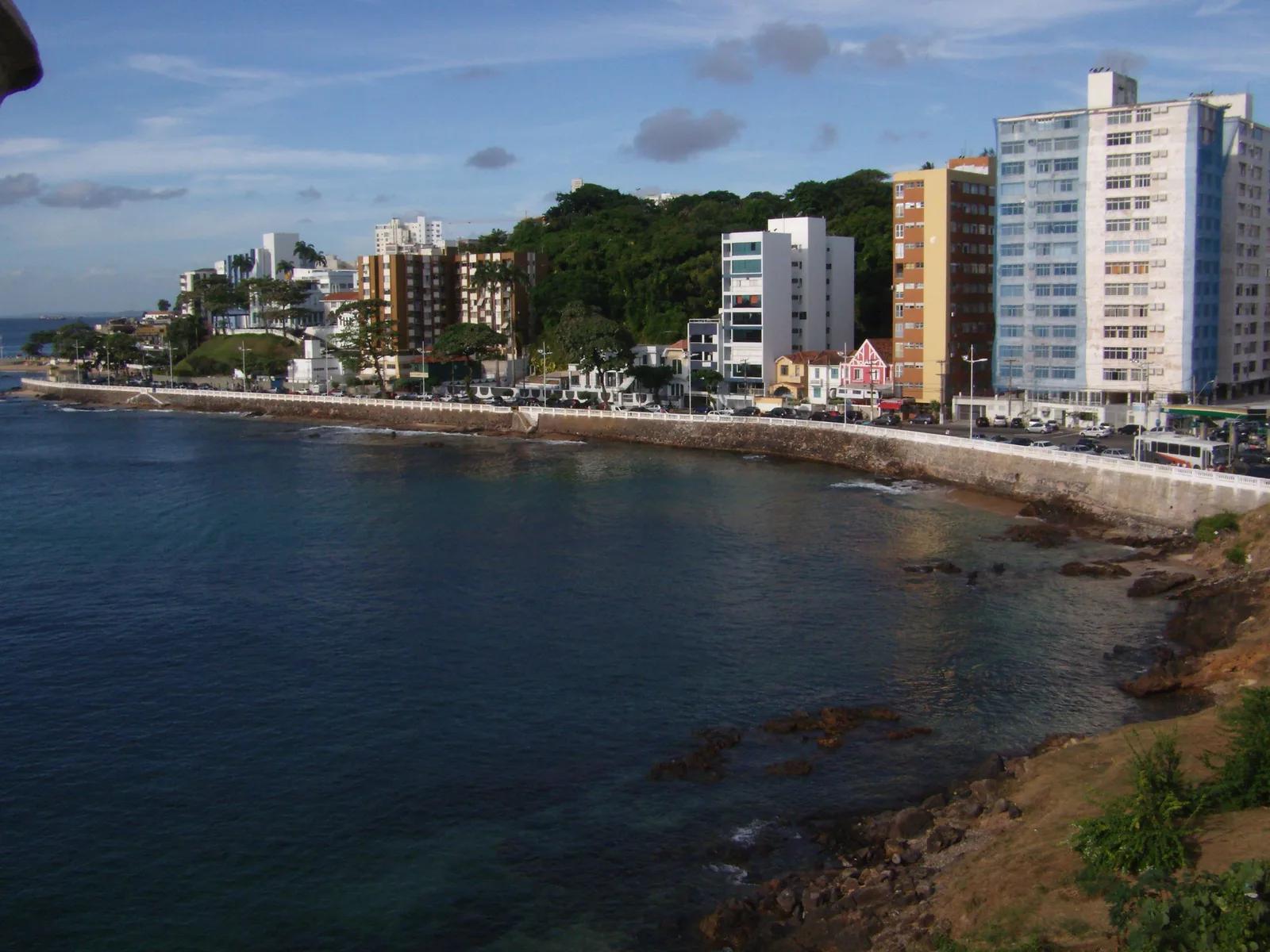 The seafront north of Barra seen from the fort at the point: the Avenida Oceânica curving along a rocky shore below a line of apartment blocks, with flat blue water in the foreground