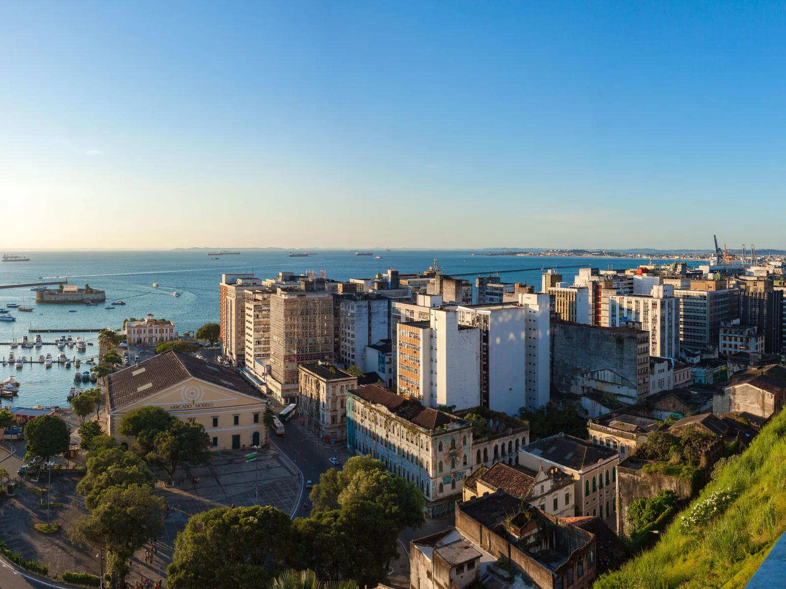 The Bay of All Saints in the late afternoon from the top of the cliff: the marina below packed with moored boats, the yellow Mercado Modelo and the office blocks of the lower city along the shore, and cargo ships out on the open water