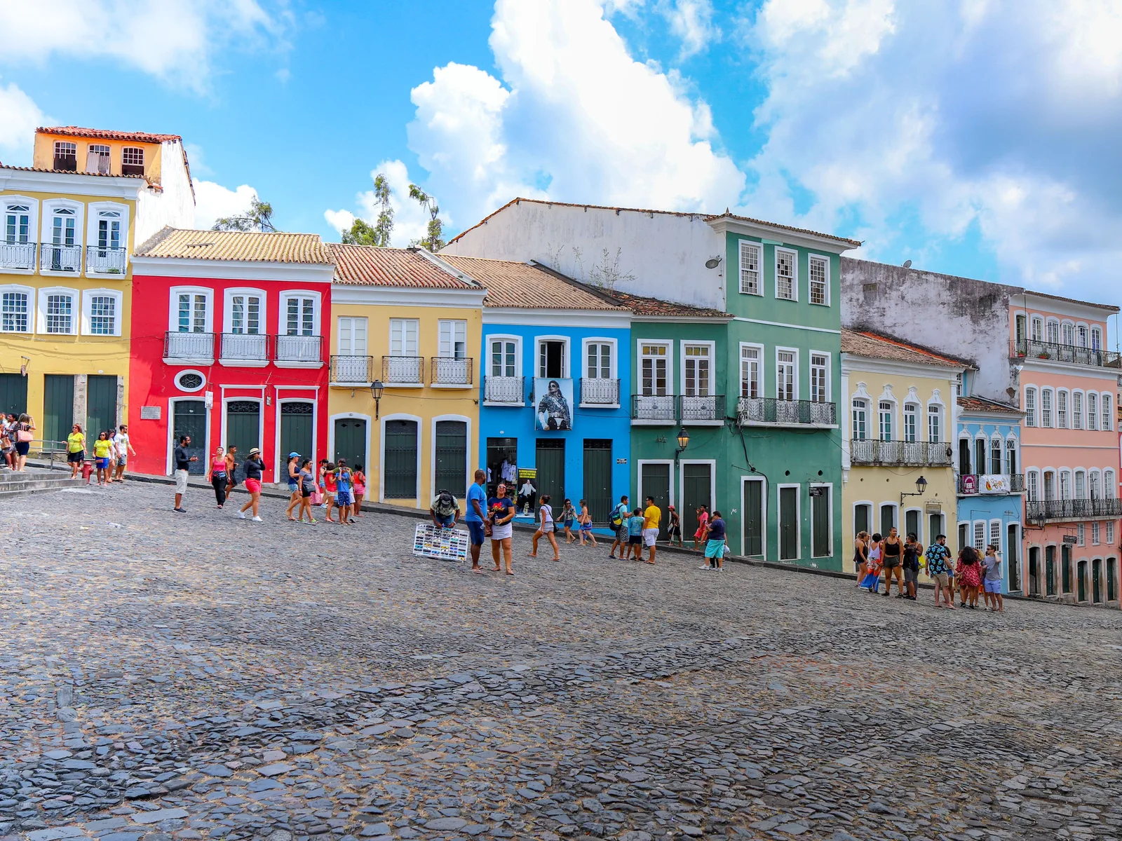 The wide cobbled expanse of the Largo do Pelourinho under a bright sky, one side lined with red, yellow, blue and green colonial townhouses, visitors crossing the stones in front of them