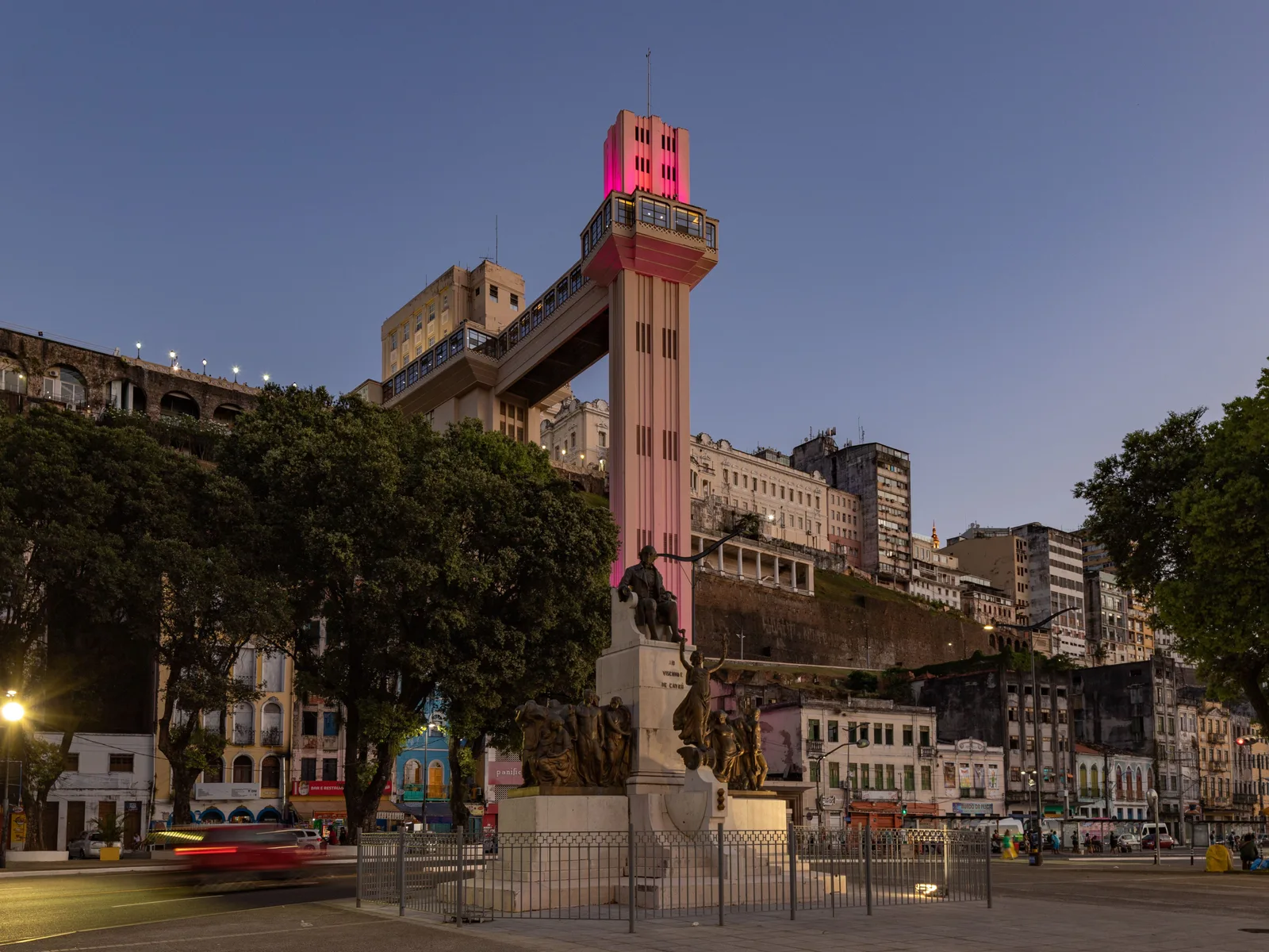The Elevador Lacerda at dusk from the Cidade Baixa below, its shafts rising the full height of the cliff to the walkway and the upper city, the tower floodlit pink for a public health campaign and traffic passing the monument at its foot