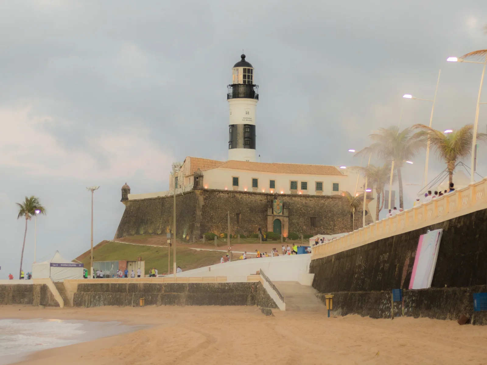 The black-and-white striped Farol da Barra lighthouse standing above the stone walls of its fort at the point, seen from the sand on an overcast evening with palms and the sea wall in the foreground