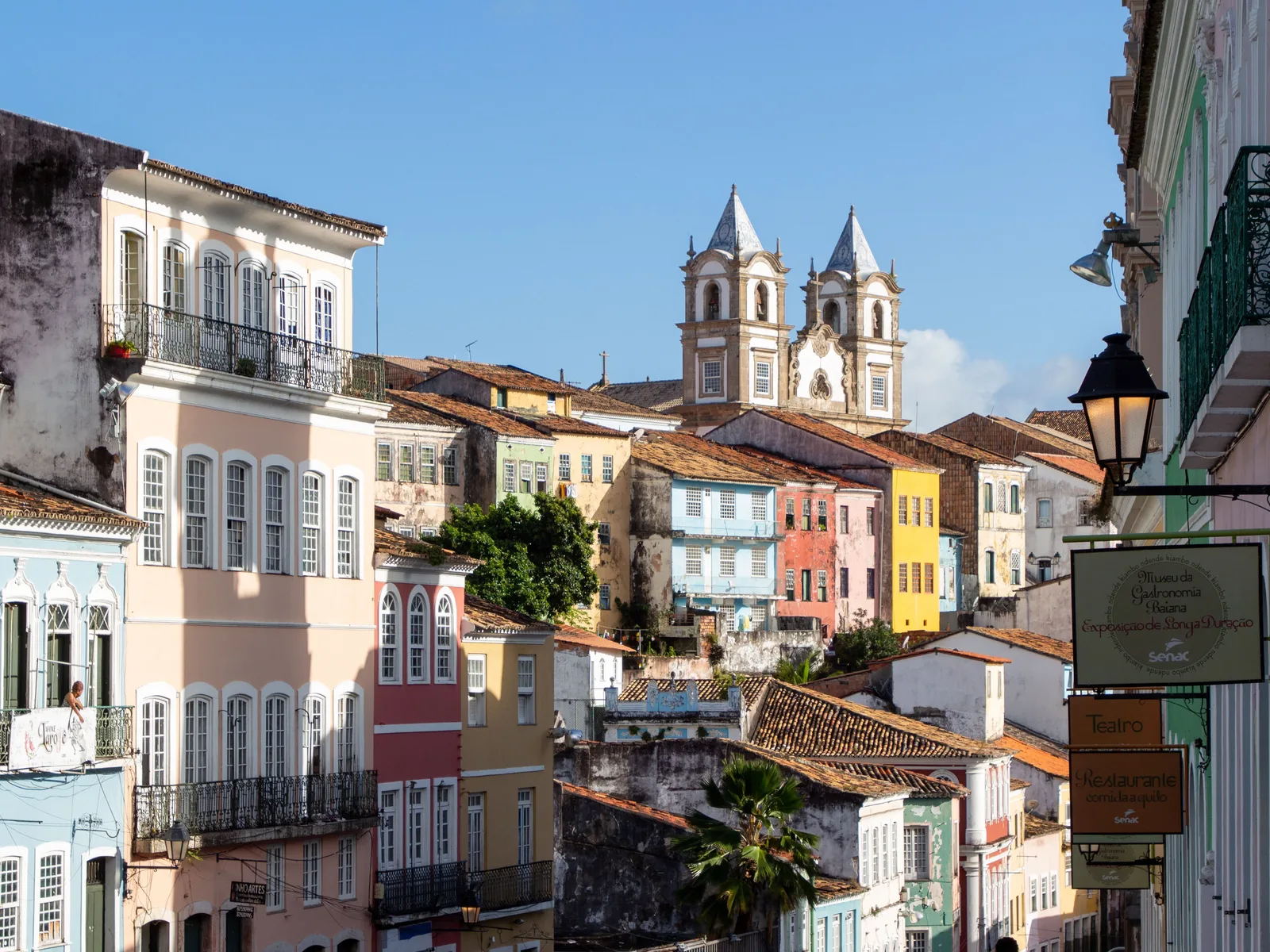 The Largo do Pelourinho from the top of the slope: rows of tall pink, blue and yellow colonial townhouses with iron balconies falling away downhill, and the twin white towers of the Rosário dos Pretos church rising over the rooftops at the bottom