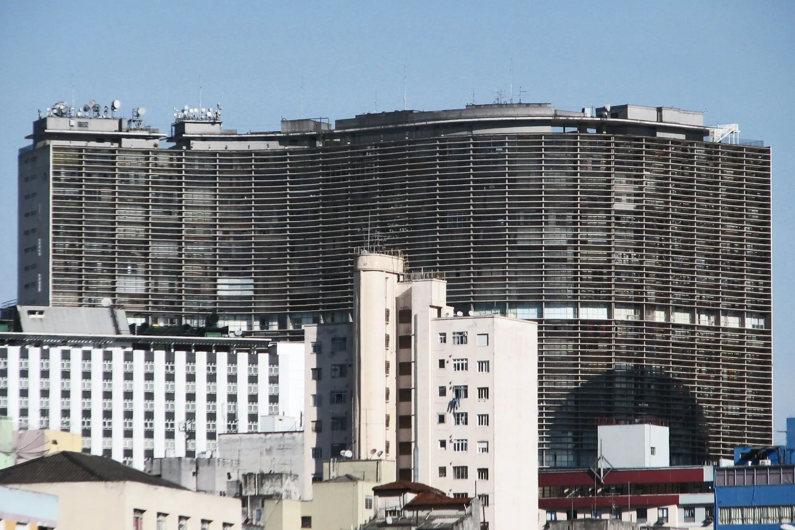 The Copan building seen from a distance over the rooftops of central São Paulo, its long curving slab banded top to bottom with concrete brise-soleil, rising dark against a clear sky behind a huddle of lower white apartment blocks