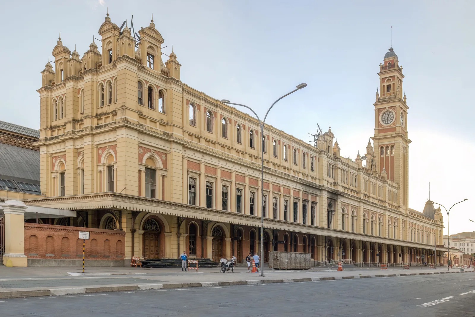 The Estação da Luz across an empty street: a long Victorian station front in cream stucco and red brick, arched windows above an iron canopy running the length of the pavement, and its tall clock tower standing at the far end under a pale hazy sky