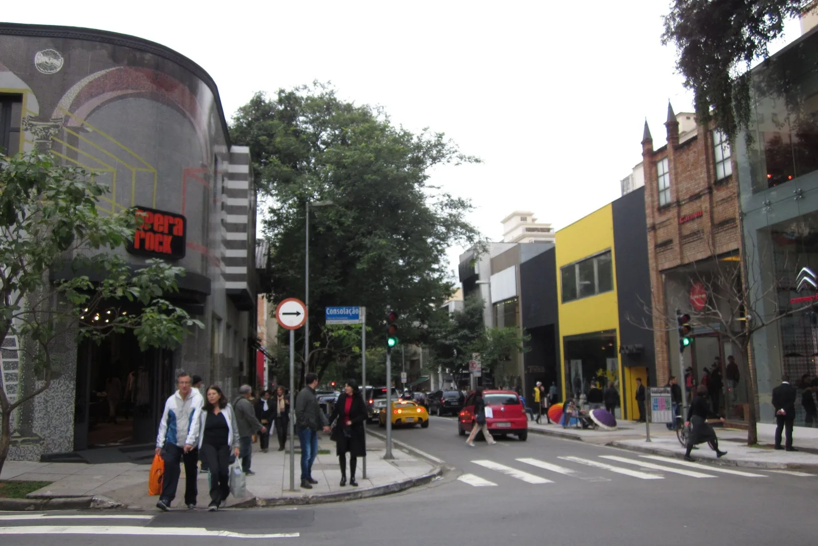 Rua Oscar Freire in Jardins on a weekday: pedestrians crossing at a marked crosswalk between low shopfronts and street trees, with a yellow building and traffic lights along the block