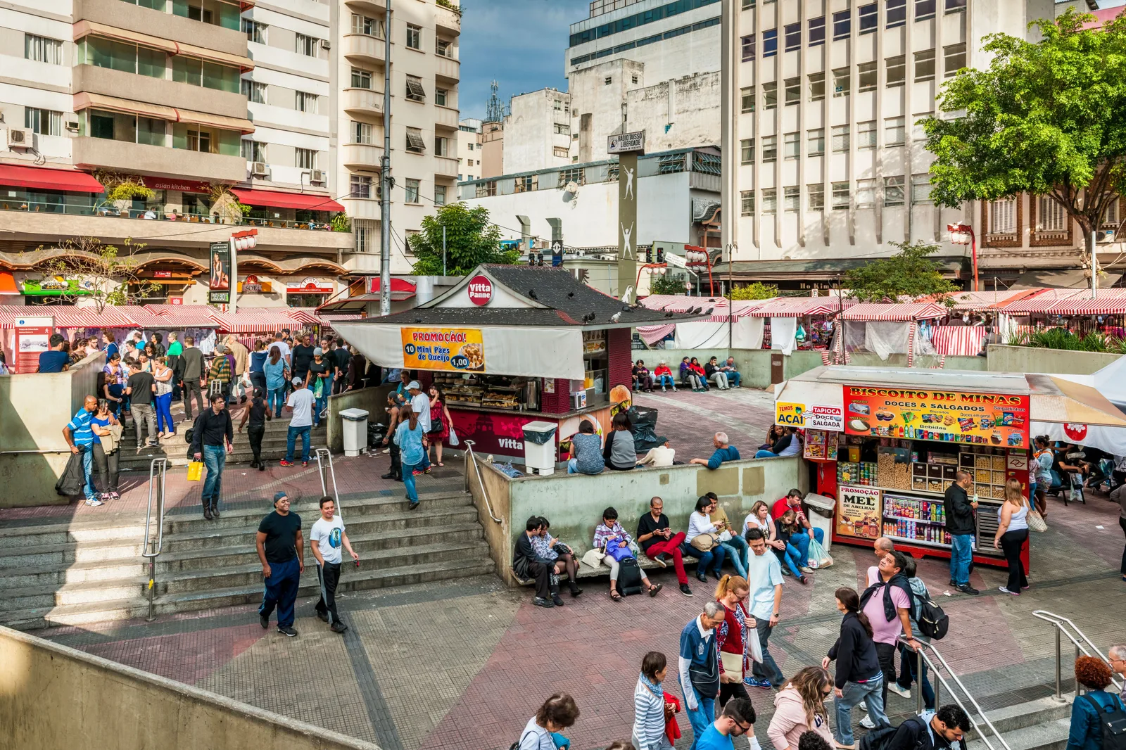 The Praça da Liberdade filled with the weekend fair: rows of red-and-white striped stall canopies down both sides, a crowd crossing the paved square and coming up the steps out of the metro, a kiosk with a black Japanese-style tiled roof in the middle of it and apartment blocks rising behind