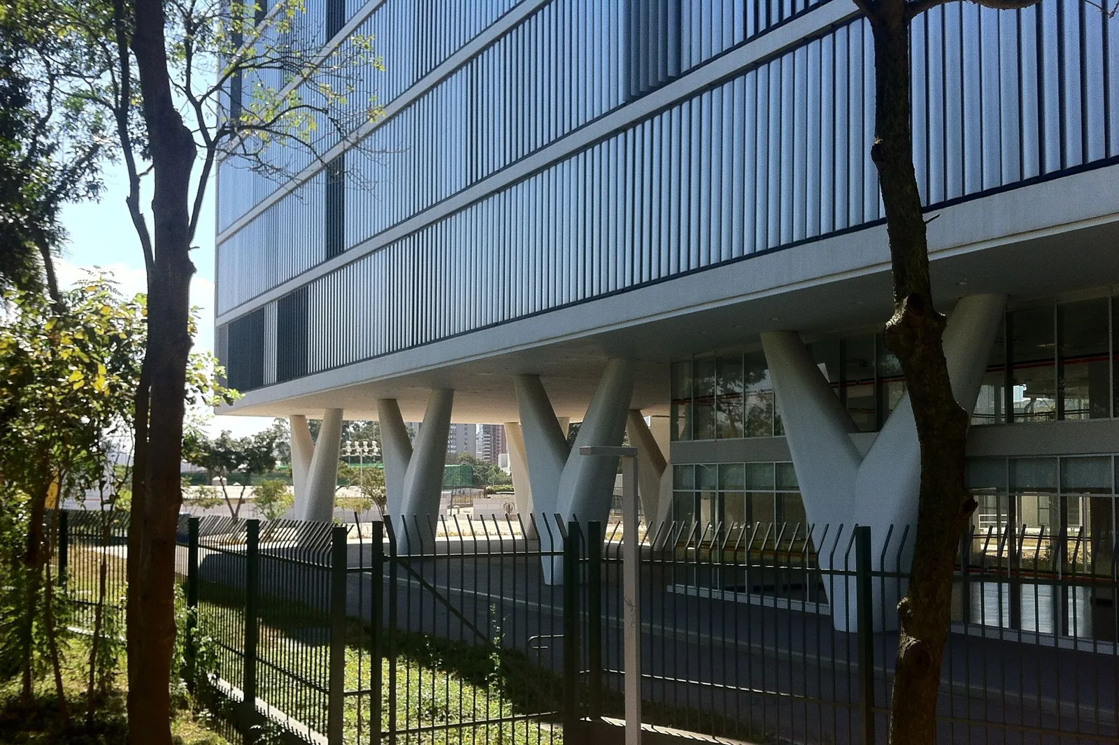 The MAC USP building beside Ibirapuera: a long horizontal block of glass and louvres raised on splayed white concrete supports, seen through park railings and bare trees