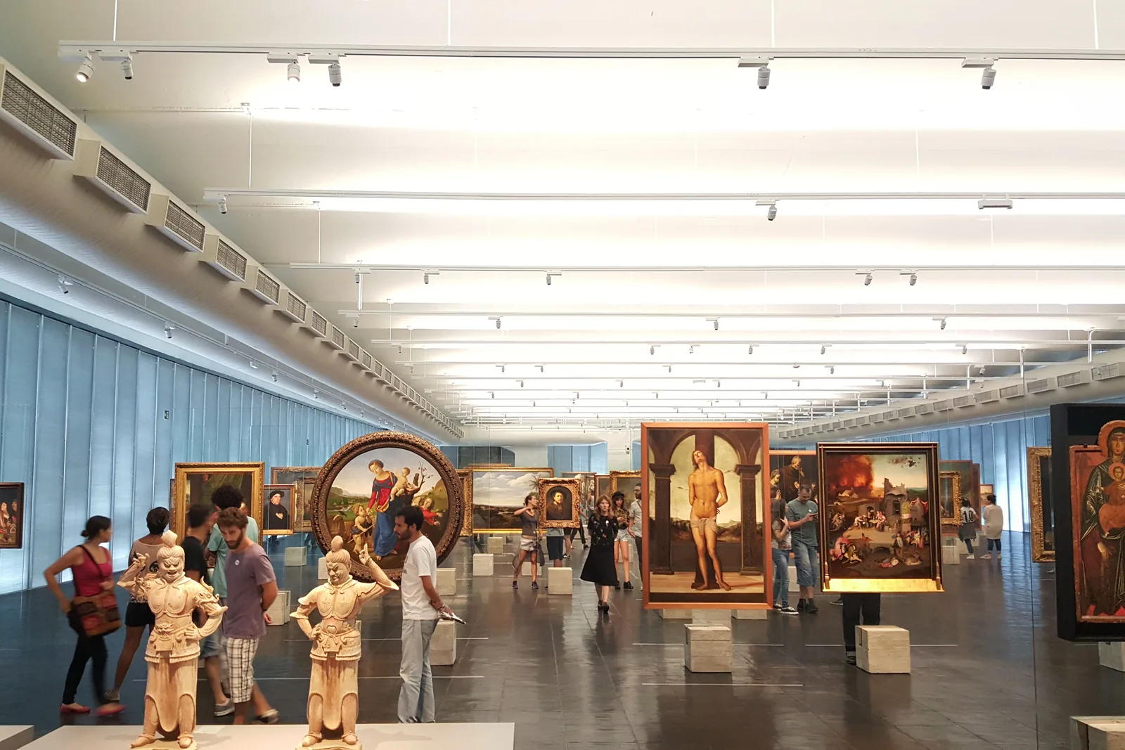 The picture gallery inside MASP: dozens of paintings mounted on freestanding glass easels standing in loose rows across one open hall with a dark polished floor and a long strip-lit ceiling, pale blue glass walls at the sides, and visitors walking between the pictures