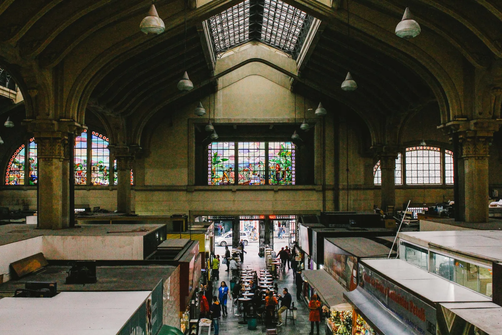 The interior of the Mercado Municipal from the mezzanine: a high vaulted hall with tall stained-glass windows at the far end, rows of stalls on the floor below and shoppers moving between them