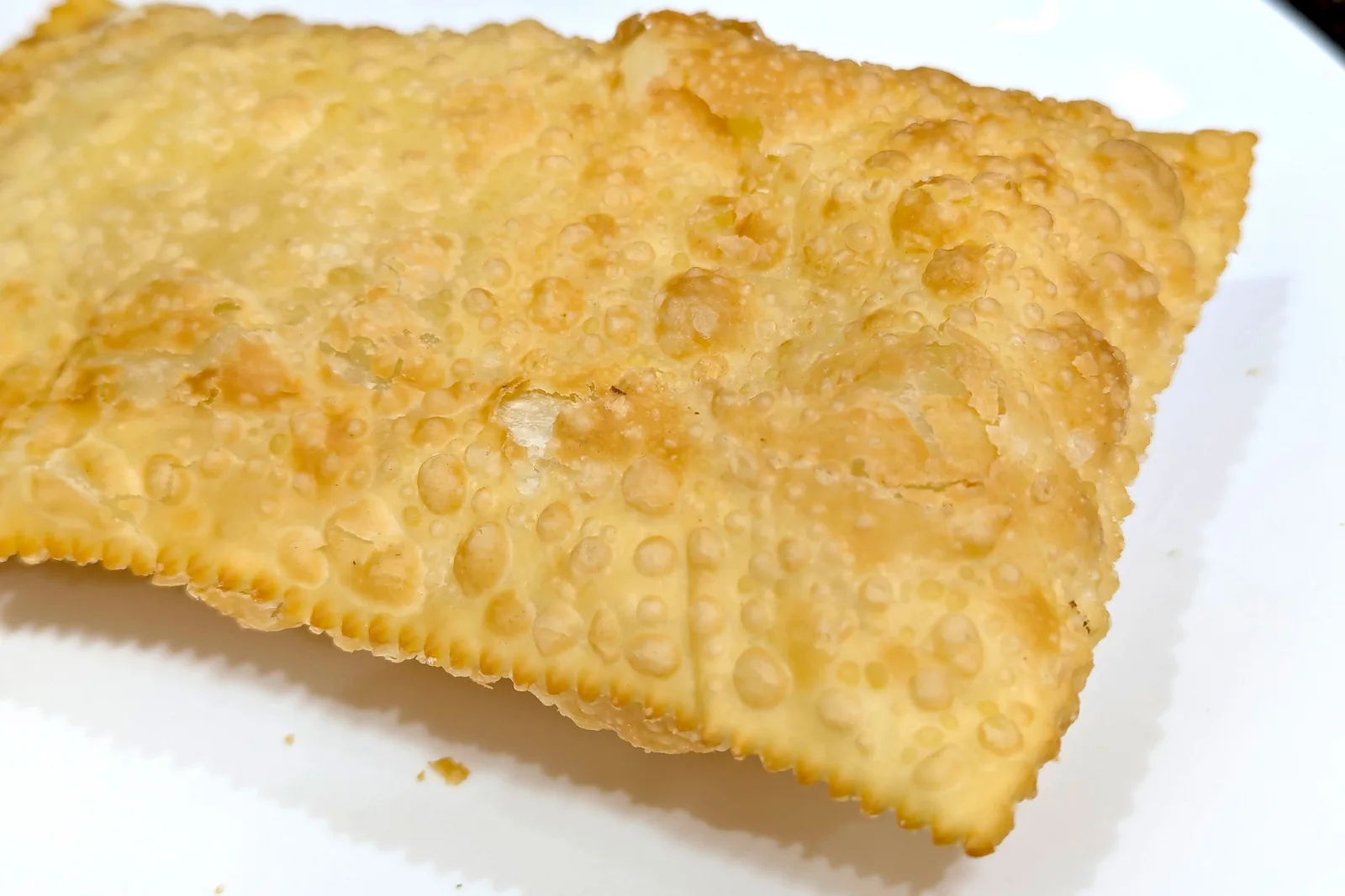 A large golden fried pastel filling a white plate, its pastry envelope blistered all over from the fryer and crimped along the edges