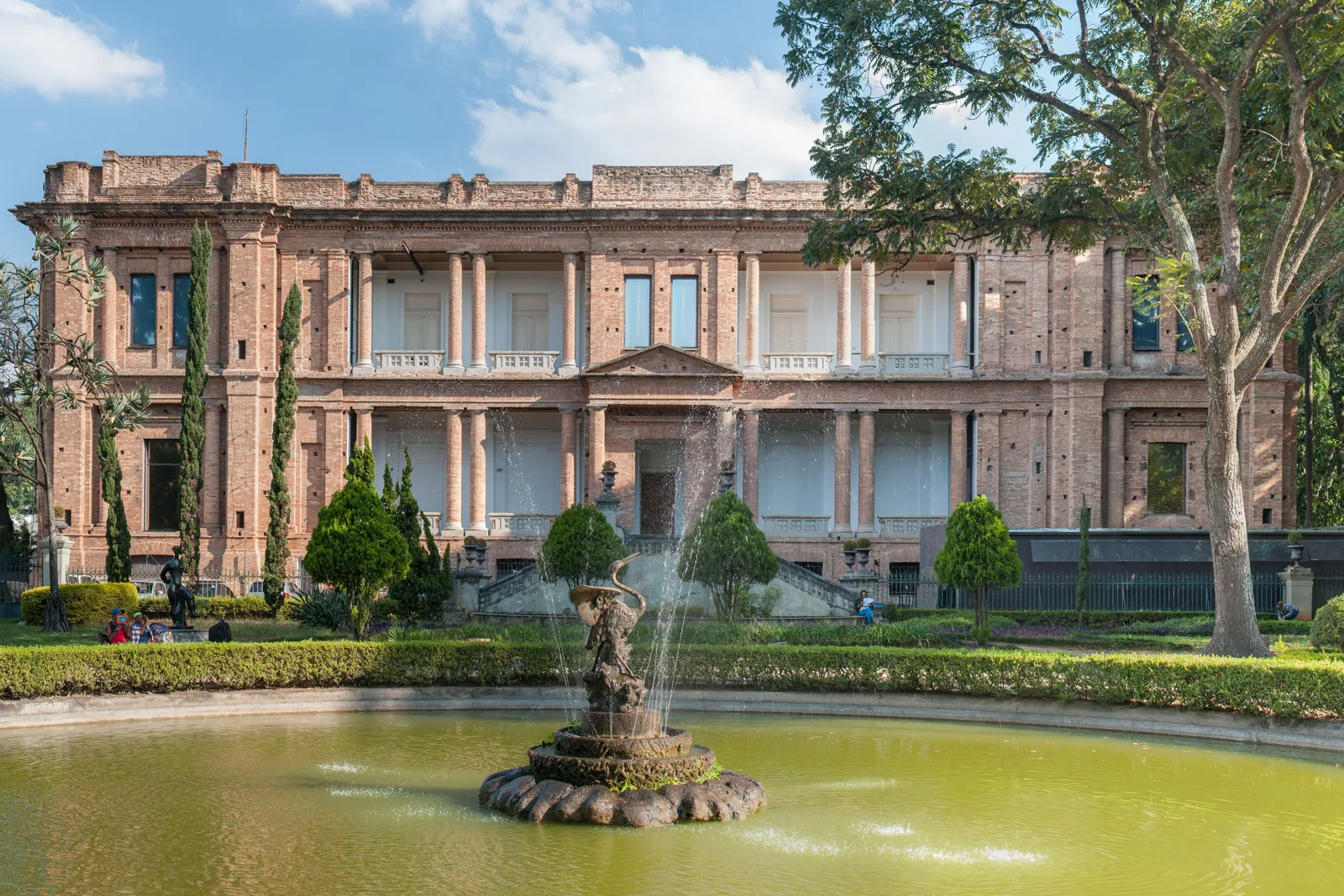 The Pinacoteca’s long bare-brick facade — two stories of brick pilasters, balustrades and tall rectangular windows — seen from the Jardim da Luz across an oval ornamental pond with a stone fountain playing in the middle of it
