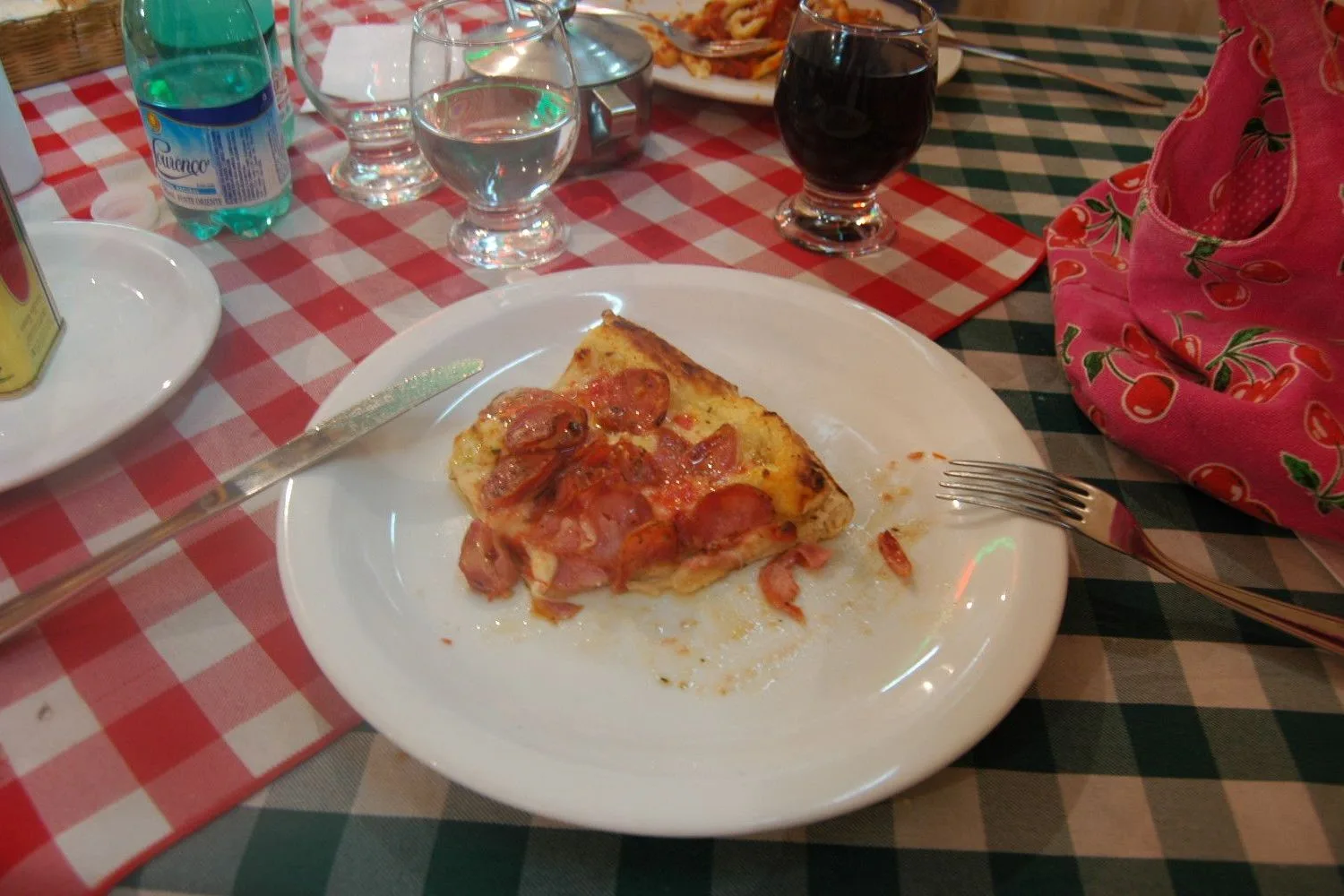A slice of tomato-topped pizza on a white plate at a pizzeria table laid with a red checked cloth, a glass of red wine and a red napkin holder beside it