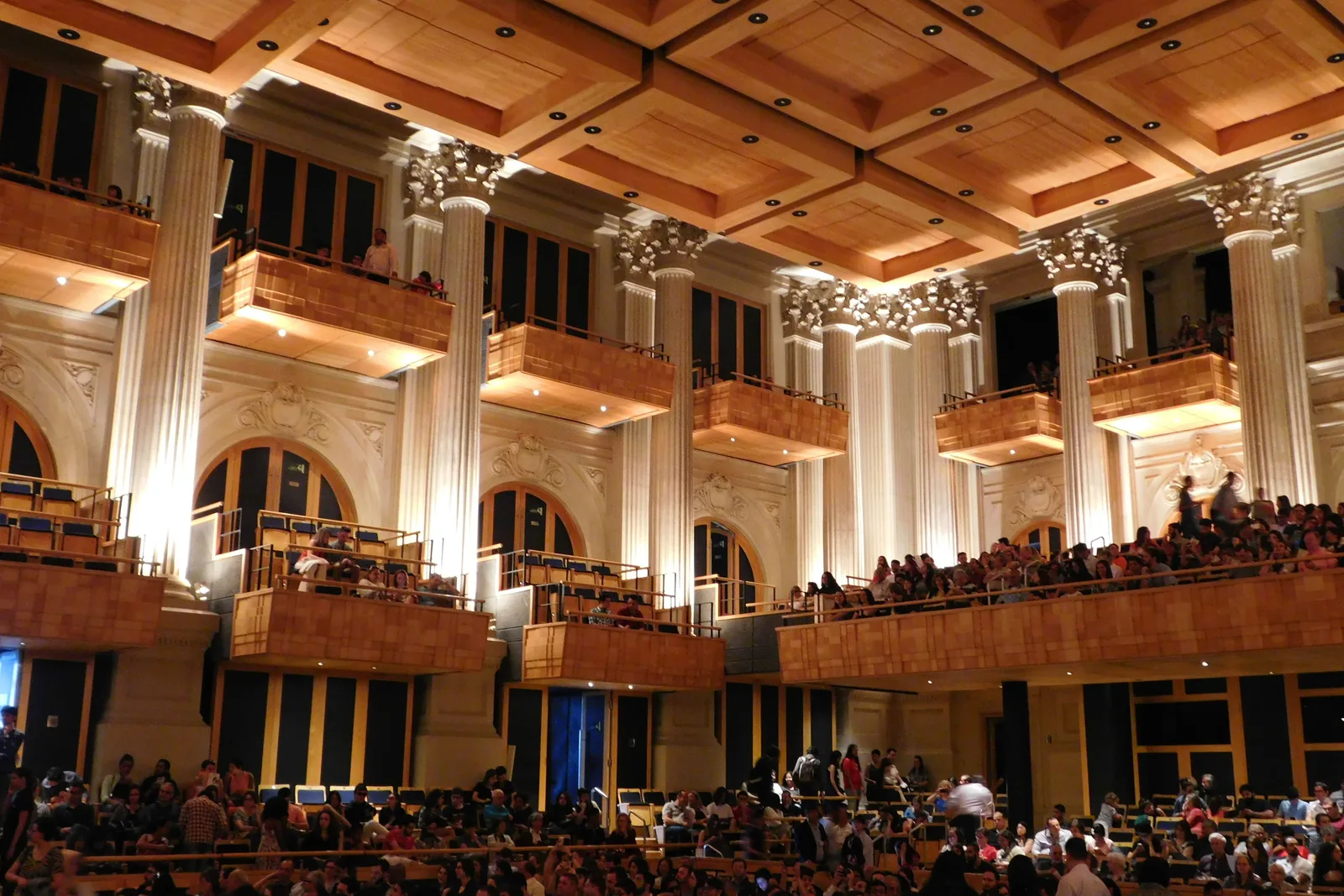 The interior of Sala São Paulo: a tall hall with cream classical walls and fluted Corinthian columns, two tiers of pale wooden boxes down the side, a deep coffered wooden ceiling overhead and the audience filling the boxes and the stalls below