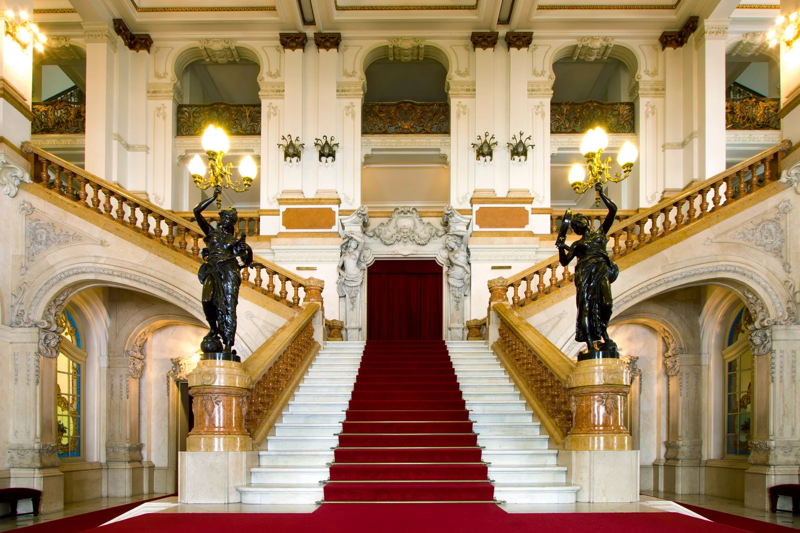 The grand staircase inside the Theatro Municipal: a red carpet running up white marble steps between gilded arches, bronze figures holding lamps on the newel posts and chandeliers overhead