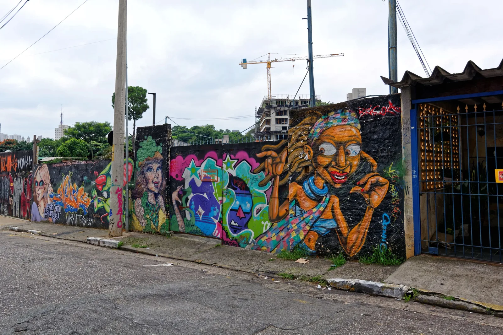 A Vila Madalena street corner with a long wall of bright graffiti murals in blue, pink and green running down the block, low buildings and a construction crane behind