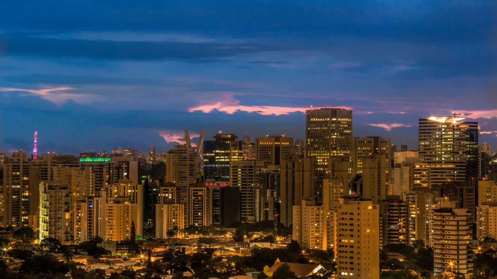 São Paulo at blue hour from above: a field of lit apartment towers running to the horizon under a deep blue sky streaked with pink cloud, office windows and street lights glowing across the whole frame