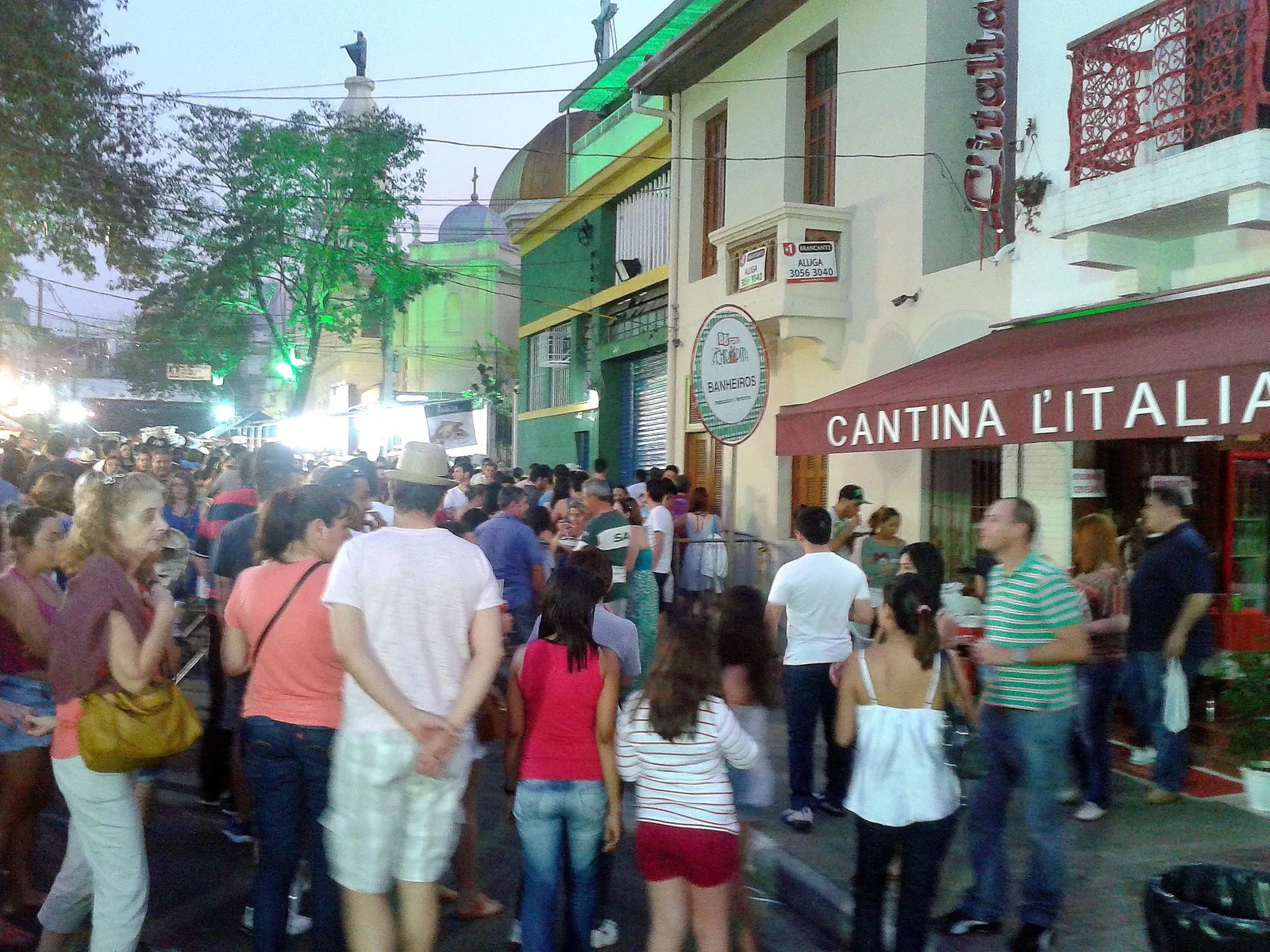 A Bixiga street at dusk during the Festa da Achiropita, packed shoulder to shoulder with people, the red awning and neon sign of the Cantina L’Italia along one side, the lit stalls of the festa receding up the street and the dome of the Achiropita church floodlit green at the end of it