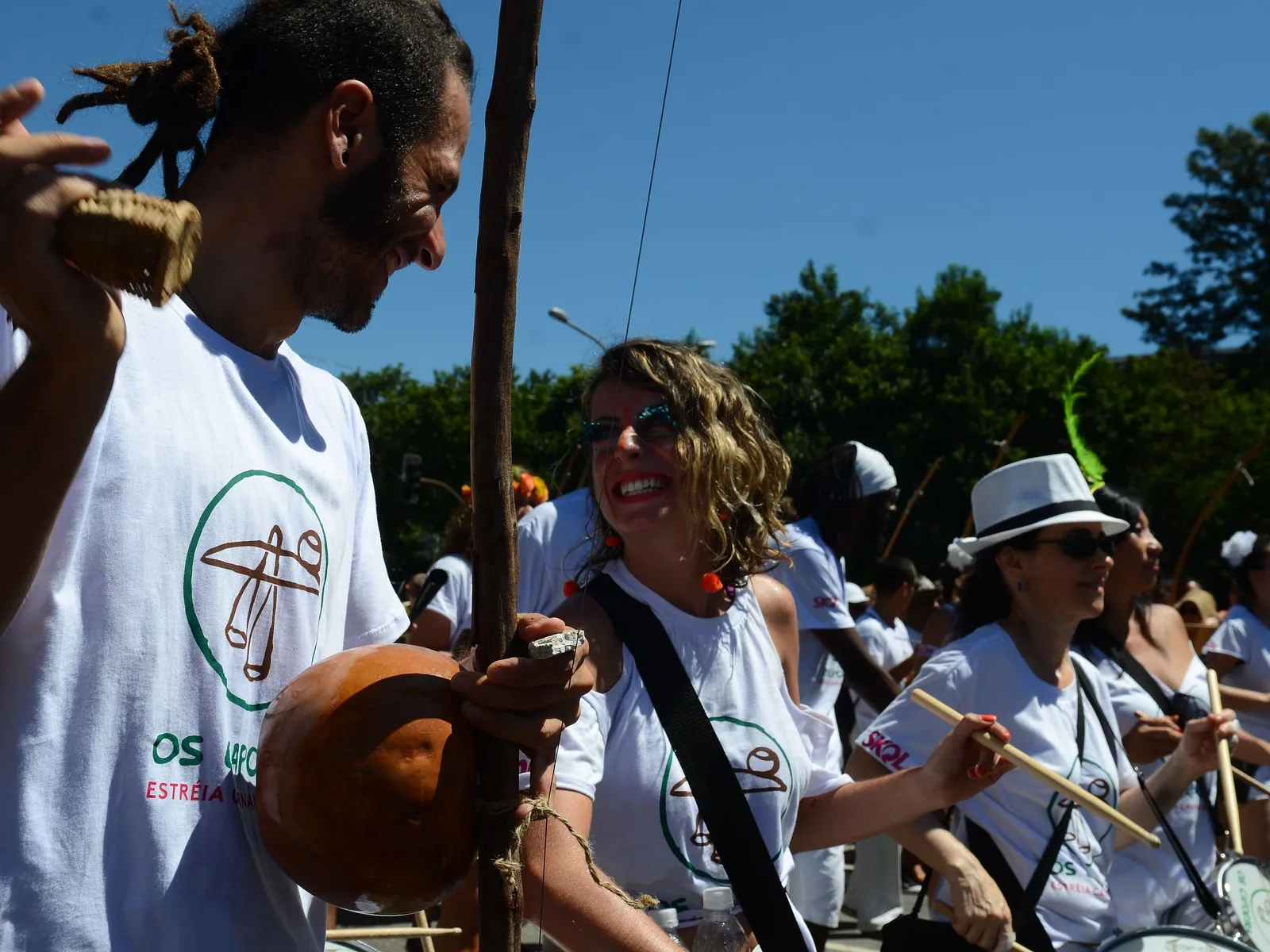Members of a São Paulo street bloco in matching white printed t-shirts and straw hats moving along a sunlit avenue, carrying drums and drumsticks, laughing as they walk