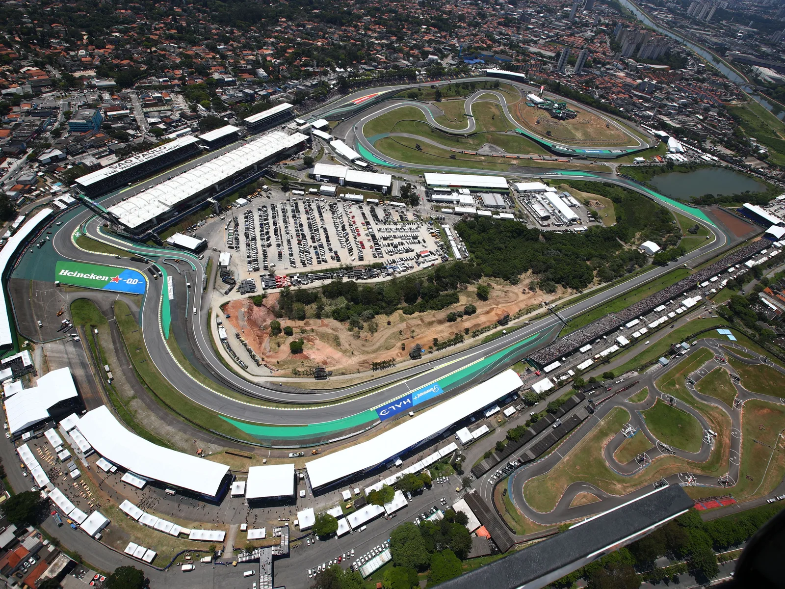 The Interlagos circuit from the air: the tight loop of gray asphalt track with green run-off and grandstands, the pit straight and paddock buildings in the middle, and the houses of southern São Paulo pressing right up to the fences
