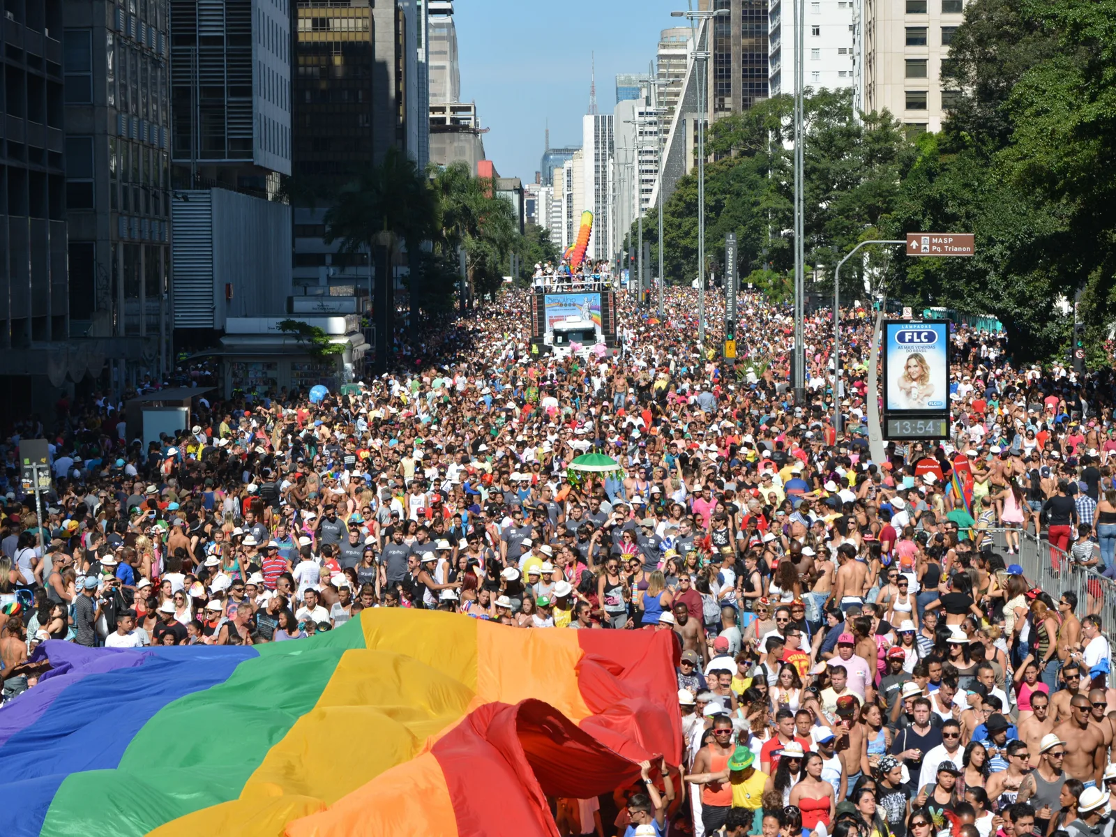 Avenida Paulista packed shoulder to shoulder with an enormous crowd for the Pride parade, a huge rainbow flag stretched across the road in the foreground and sound trucks and towers receding into the distance