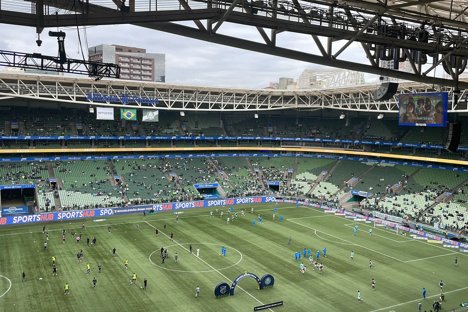 Palmeiras’s arena in Água Branca seen from high in the stand before kickoff: two teams warming up across the green pitch under a flat gray sky, the green seats still half empty, advertising boards around the touchline and the stadium’s steel roof trusses cutting across the top of the frame