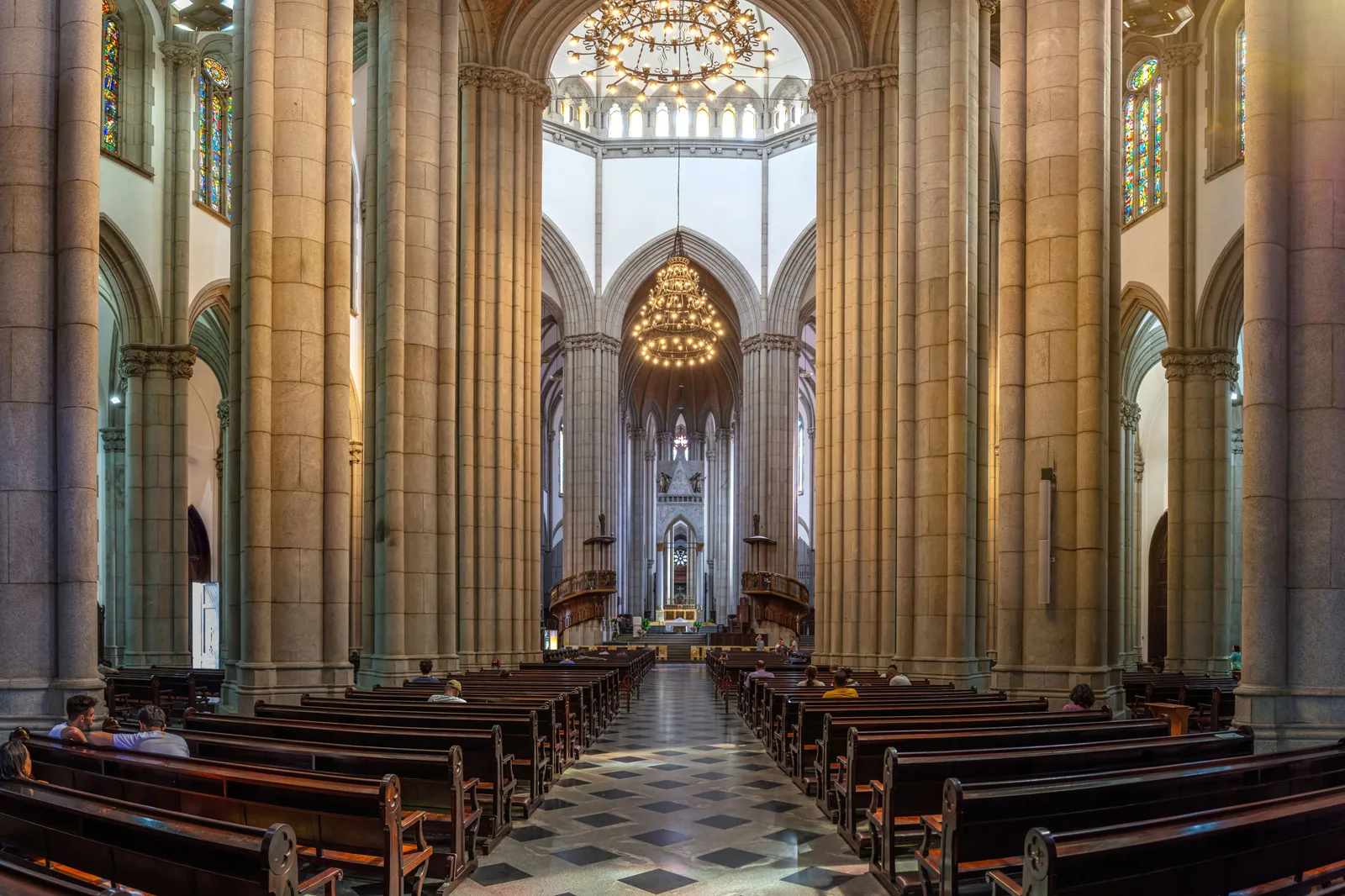 Inside the Catedral da Sé: a tall neo-Gothic nave of pale stone piers and ribbed vaults, rows of dark wooden pews along a black-and-white checkered floor, stained glass down both aisles and two large brass chandeliers hanging over the center