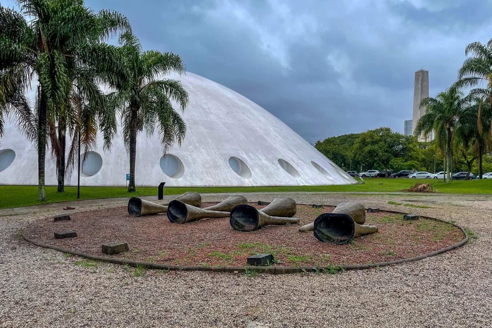 The white concrete dome of Niemeyer’s Oca pavilion in Parque Ibirapuera behind a screen of tall palms, with a ring of gravel and low sculptures in front of it