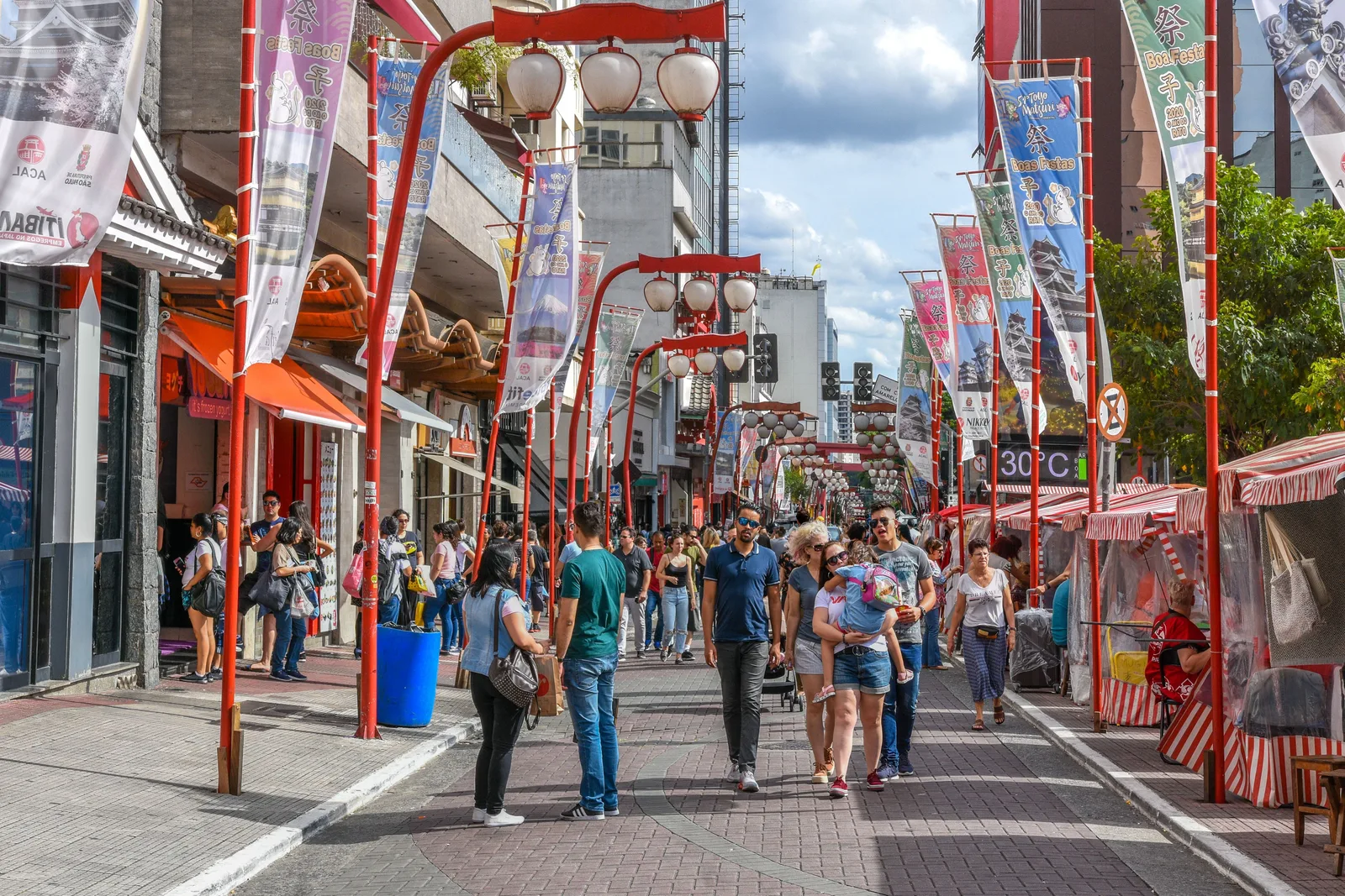 The main street of Liberdade under its red steel arches and hanging lanterns, shopfronts and banners along both sides and a crowd of shoppers filling the pavement