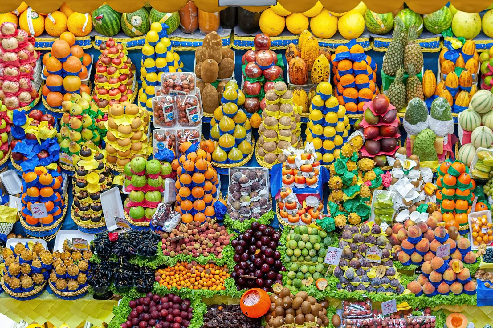 A fruit stall at the Mercado Municipal seen head-on and filling the frame: pineapples, melons, grapes, persimmons, pomegranates, dragon fruit and a dozen tropical fruits with no English name, each stacked into its own pyramid on blue and yellow paper, with handwritten price cards tucked between them