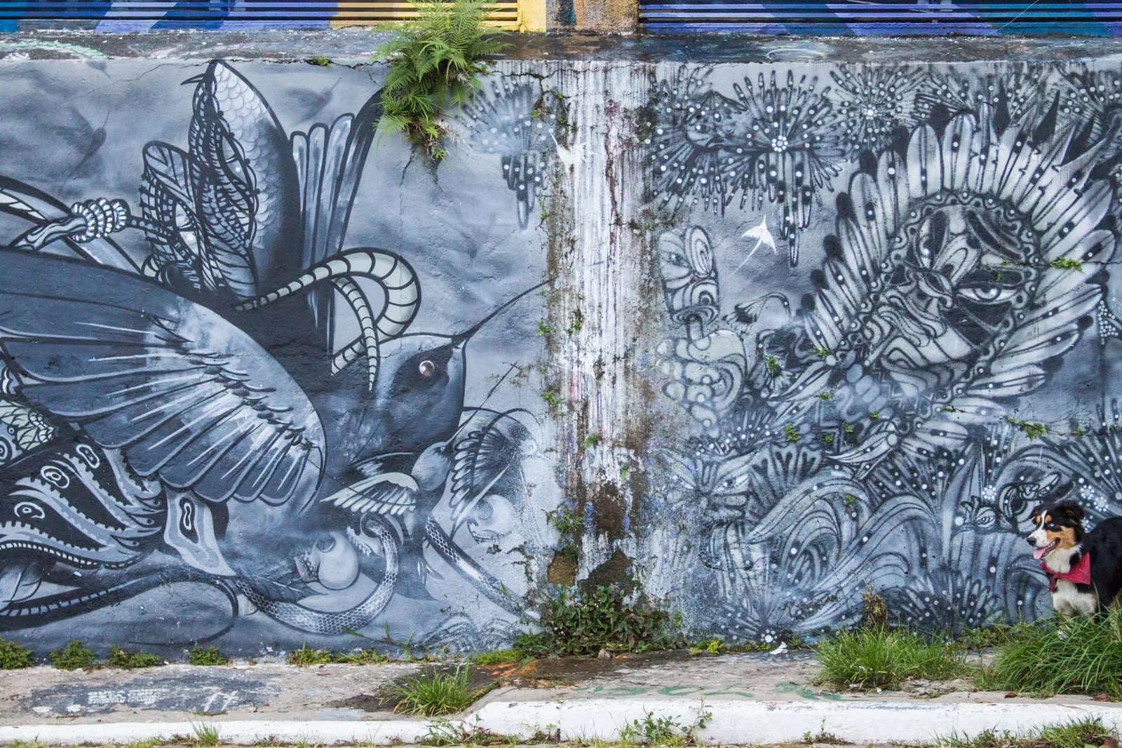 A large monochrome mural of birds and foliage covering a wall in the Beco do Batman, painted in fine gray linework, with a small figure and a strip of color at the far end