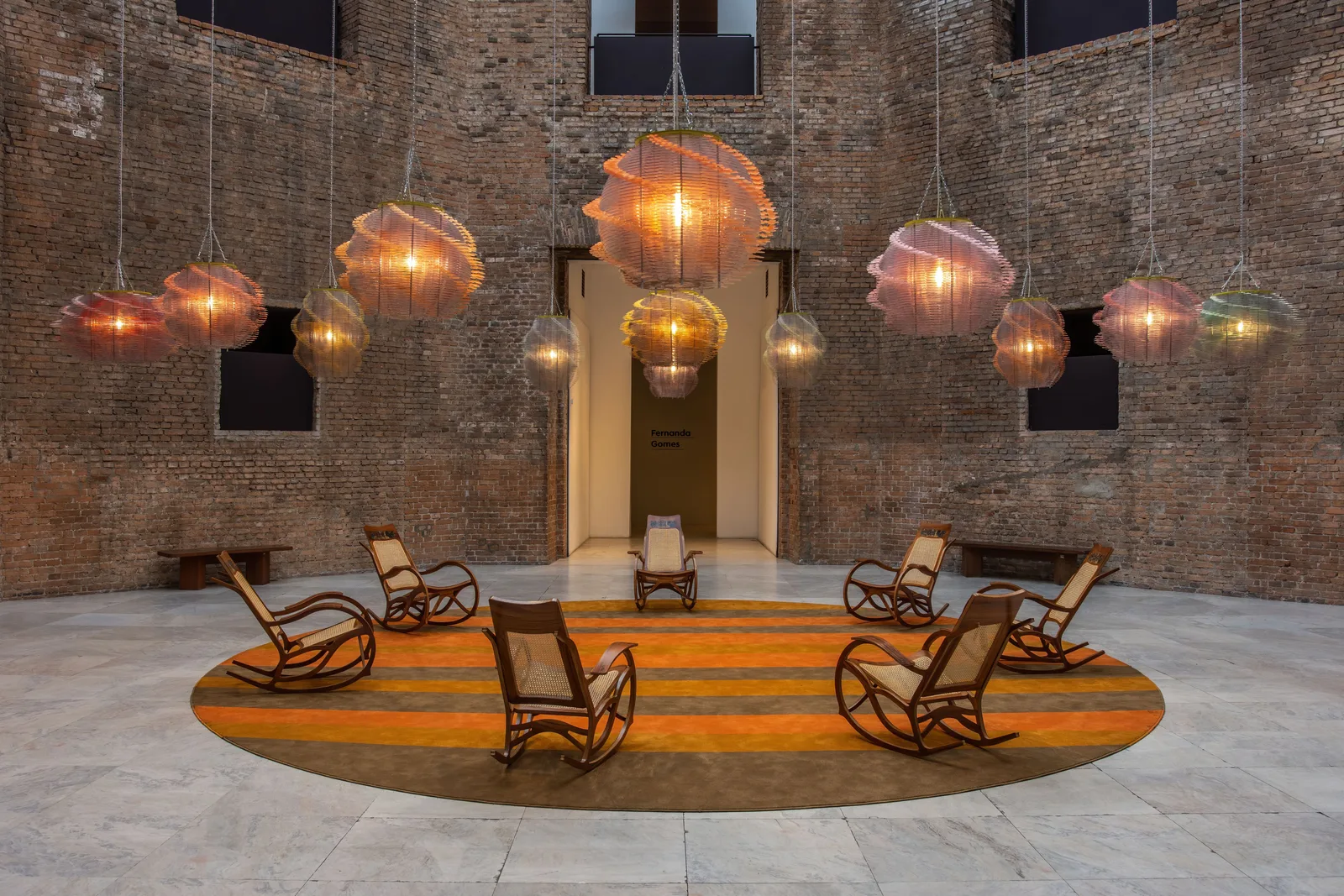 A brick hall inside the Pinacoteca with a circle of wooden rocking chairs on an orange floor under a row of hanging glass lamps